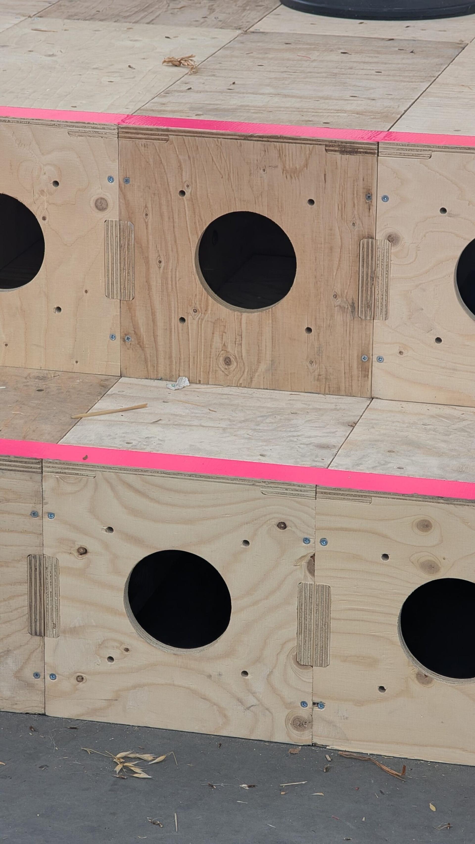 Wooden structure with multiple circular holes and bright pink tape along the edges, resembling a play or exercise area for small animals or children, crafted from sustainably sourced materials.