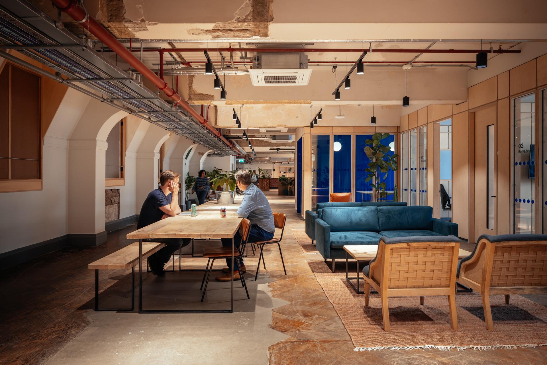Two people sit at a wooden table talking in a spacious, modern office with exposed ceilings, spruce accents, indoor plants, blue sofas, and large windows letting in natural light.