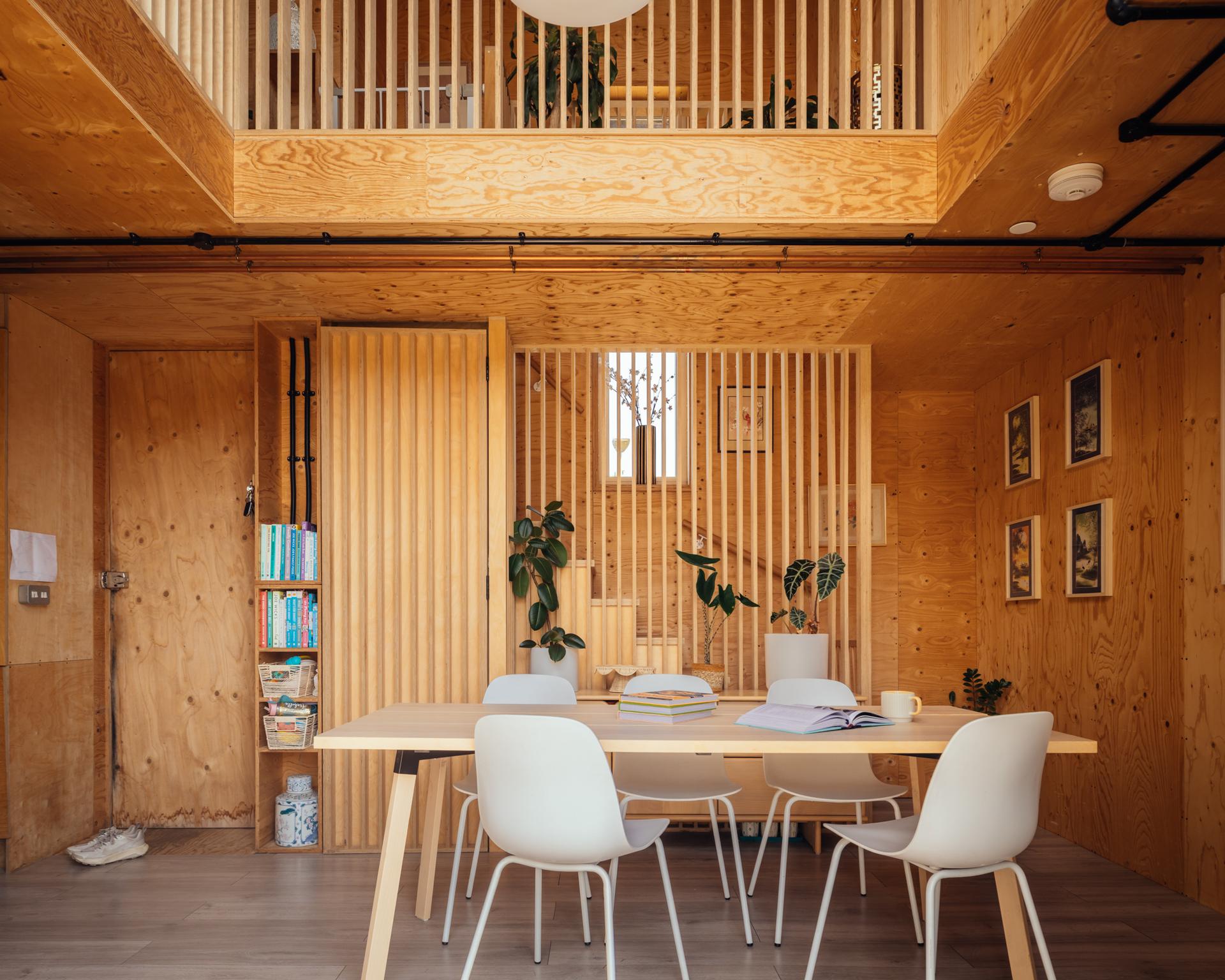 A modern home extension featuring a dining area with a light wood table and four white chairs, set in a room panelled in plywood with a slatted wooden loft overlook above.
