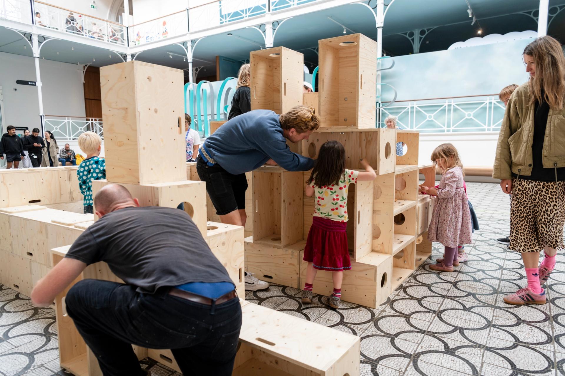 Adults and children interact and play with large U-Build wooden blocks in a spacious indoor area with a patterned floor and high ceilings. Some children are exploring the U-Build blocks while others watch or join in.