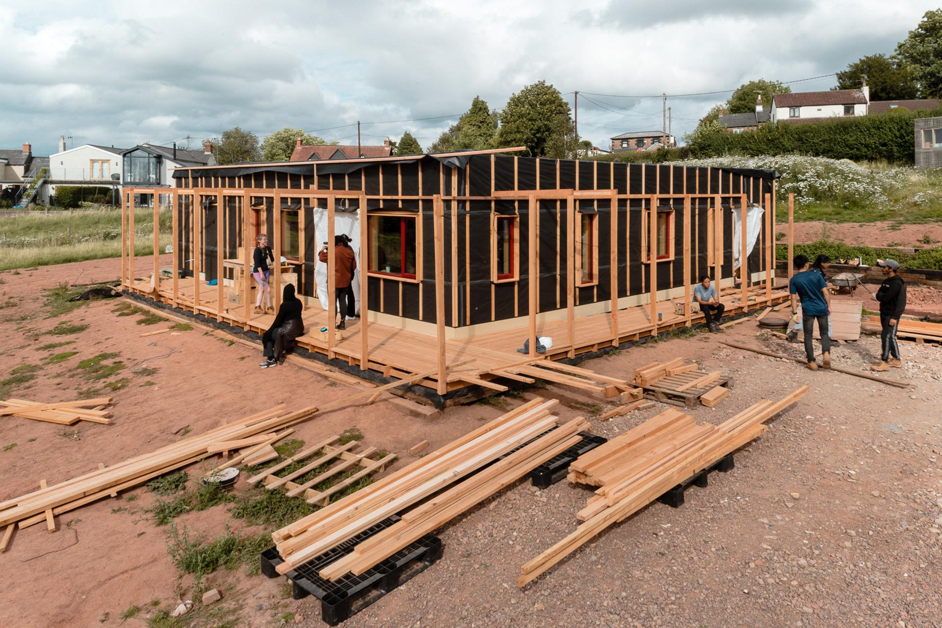 A group of people work together to construct a wooden house frame on a dirt plot, using sustainably sourced building materials and planks scattered around the site under a cloudy sky.