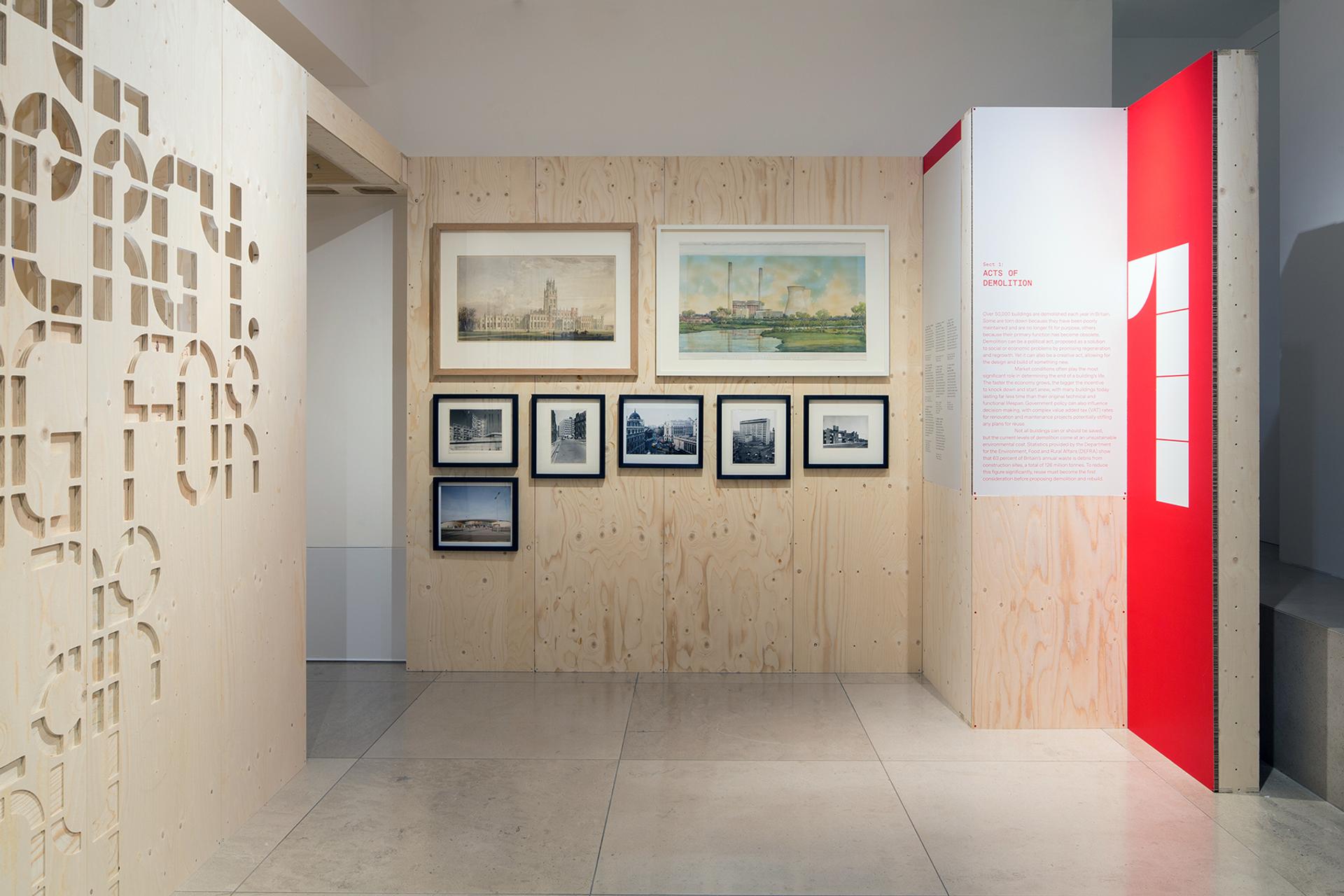 A museum exhibition features a birch wooden wall displaying six framed architectural drawings and photographs, with an adjacent red and white informational panel on the right and a decorative cut-out wooden partition on the left.