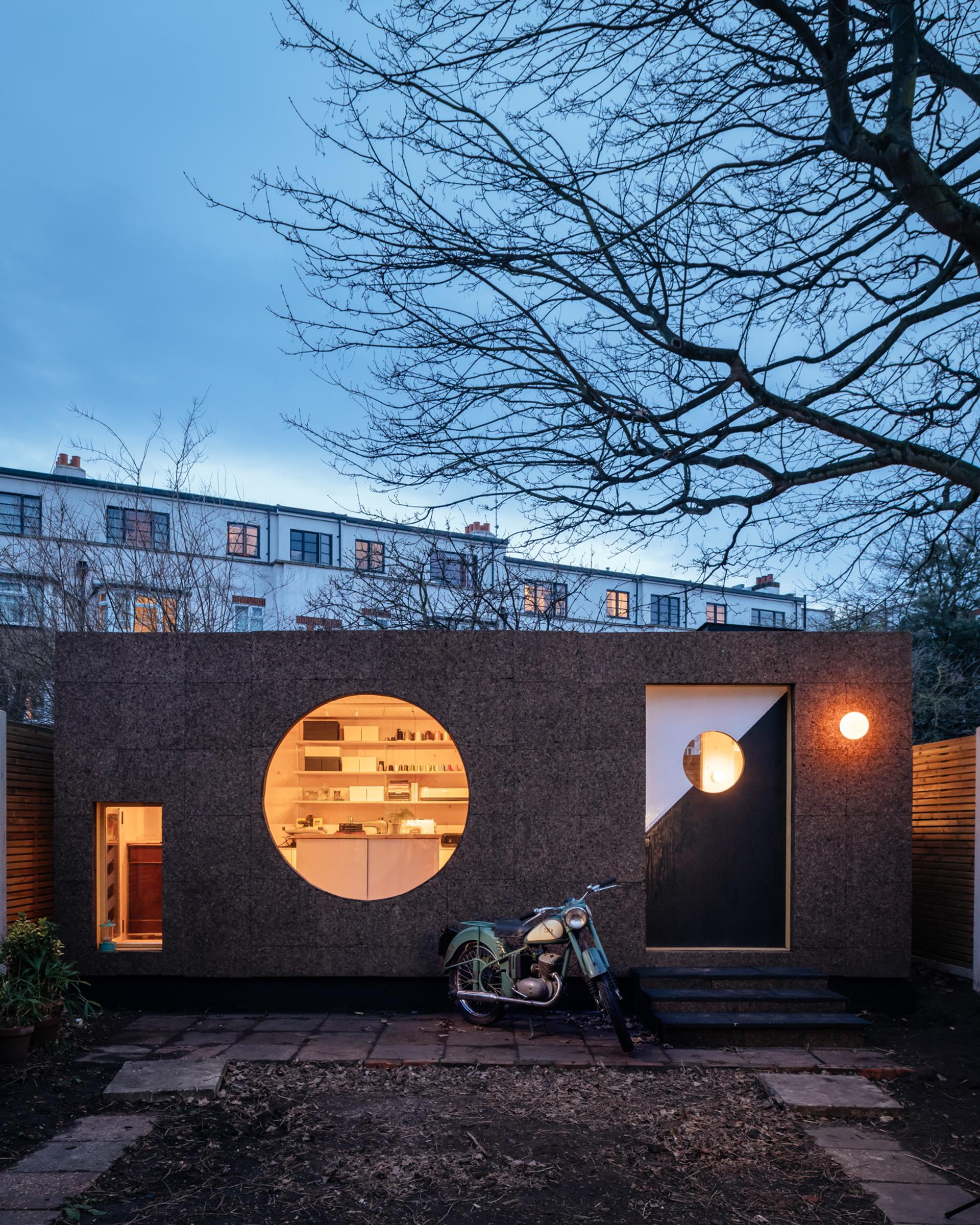 A modern, box-shaped small house with a large circular window and geometric door, warmly lit from inside, stands in a garden with a vintage motorbike parked in front—crafted with sustainably sourced materials—bare trees overhead at twilight.