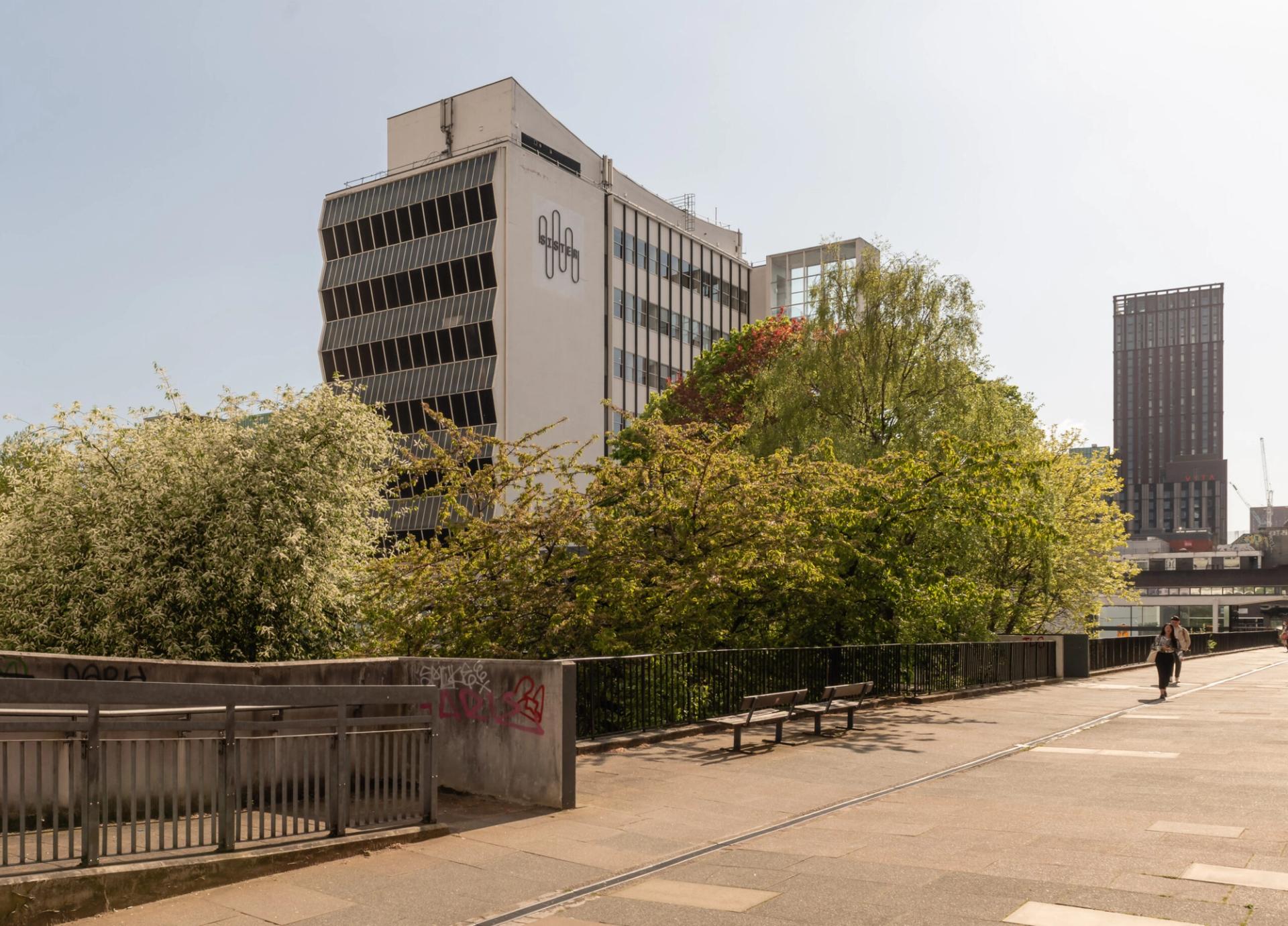 A pedestrian walks along a wide, sunny path lined with green trees and bushes in Manchester. Modern buildings, including one with a geometric logo of Sustainable Ventures, rise in the background under a clear sky.