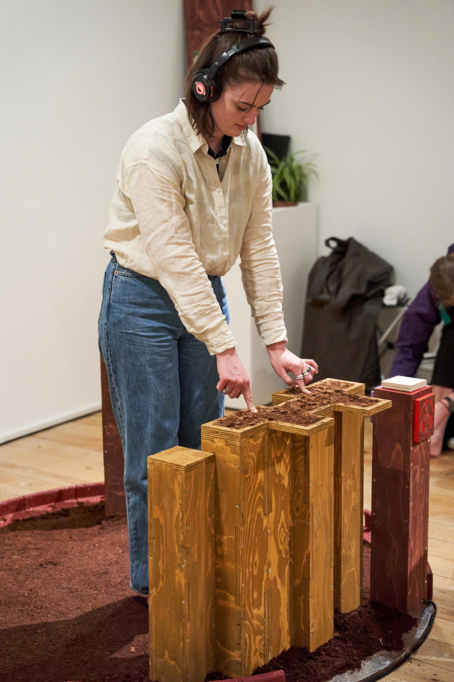 A person wearing headphones and casual clothes shapes sustainable brown material on tall, modular wooden blocks in an indoor setting, with another person bent down in the background.