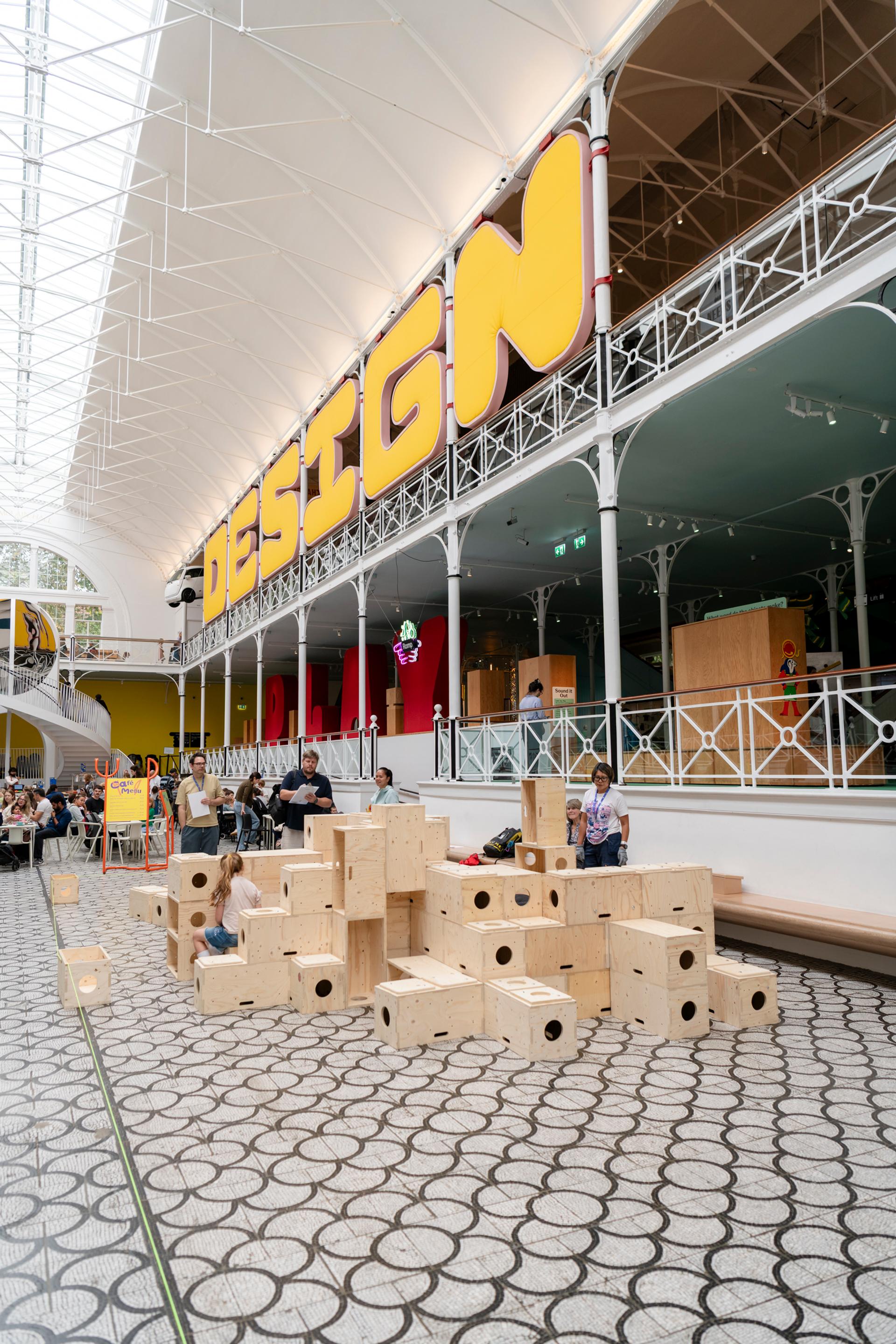 Children play on large U-Build wooden blocks in an open, modern indoor space with a high ceiling. A bold yellow DESIGN sign is displayed above a mezzanine level, and the floor features a geometric tile pattern.