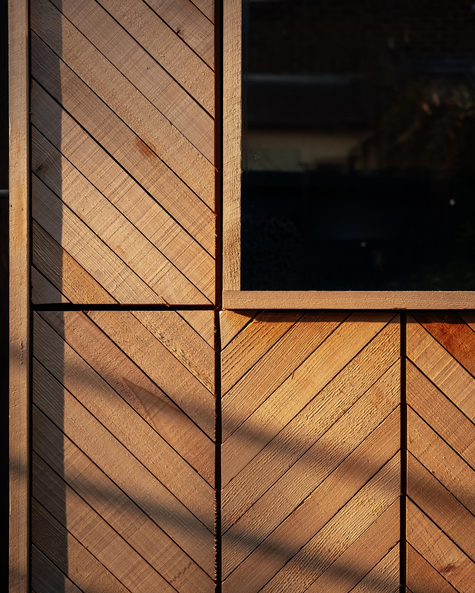 Close-up of a spruce timber construction wall with diagonal planks forming a chevron pattern, lit by sunlight, next to a dark windowpane. Shadows and light create contrast on the modular, textured surface.
