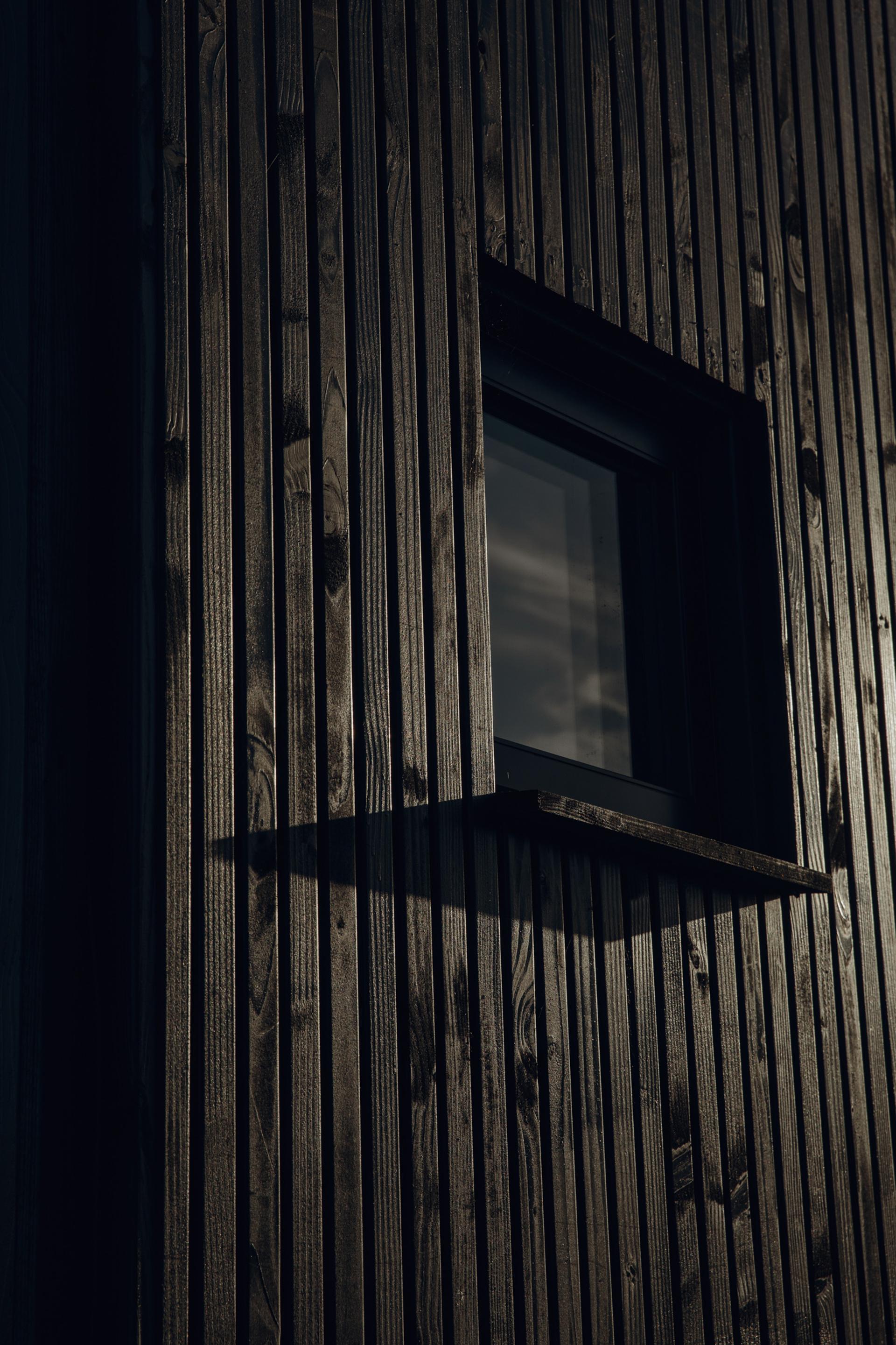 A close-up of a modern modular wooden exterior wall with vertical spruce planks, featuring a small rectangular window reflecting cloudy sky, casting sharp shadows in low, moody lighting.