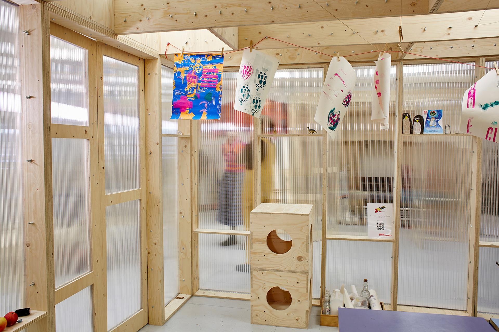 A brightly lit room with translucent plastic walls and timber construction frames. Colourful children's artwork hangs from a string, and a wooden cube structure crafted from plywood with circular cut-outs sits on the floor. Art supplies are on the shelves.