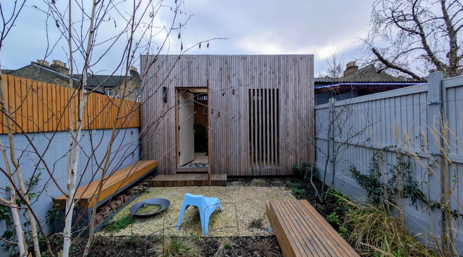A modern modular garden studio with vertical spruce timber construction, set in a small back garden with benches, gravel, a blue chair, and young trees, all surrounded by a wooden and grey fence.
