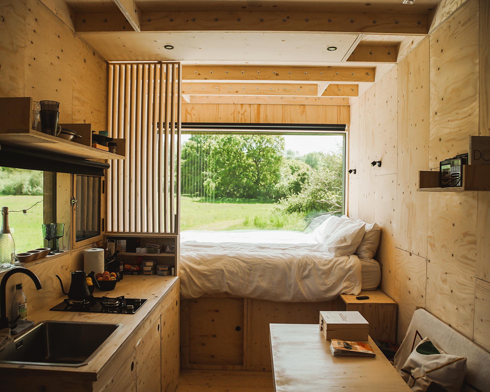 A cosy tiny house interior with light wooden walls, a small kitchen on the left, a bed with white bedding in front of a large window overlooking greenery, and a bench-style table on the right—showcasing thoughtful reuse for sustainability.