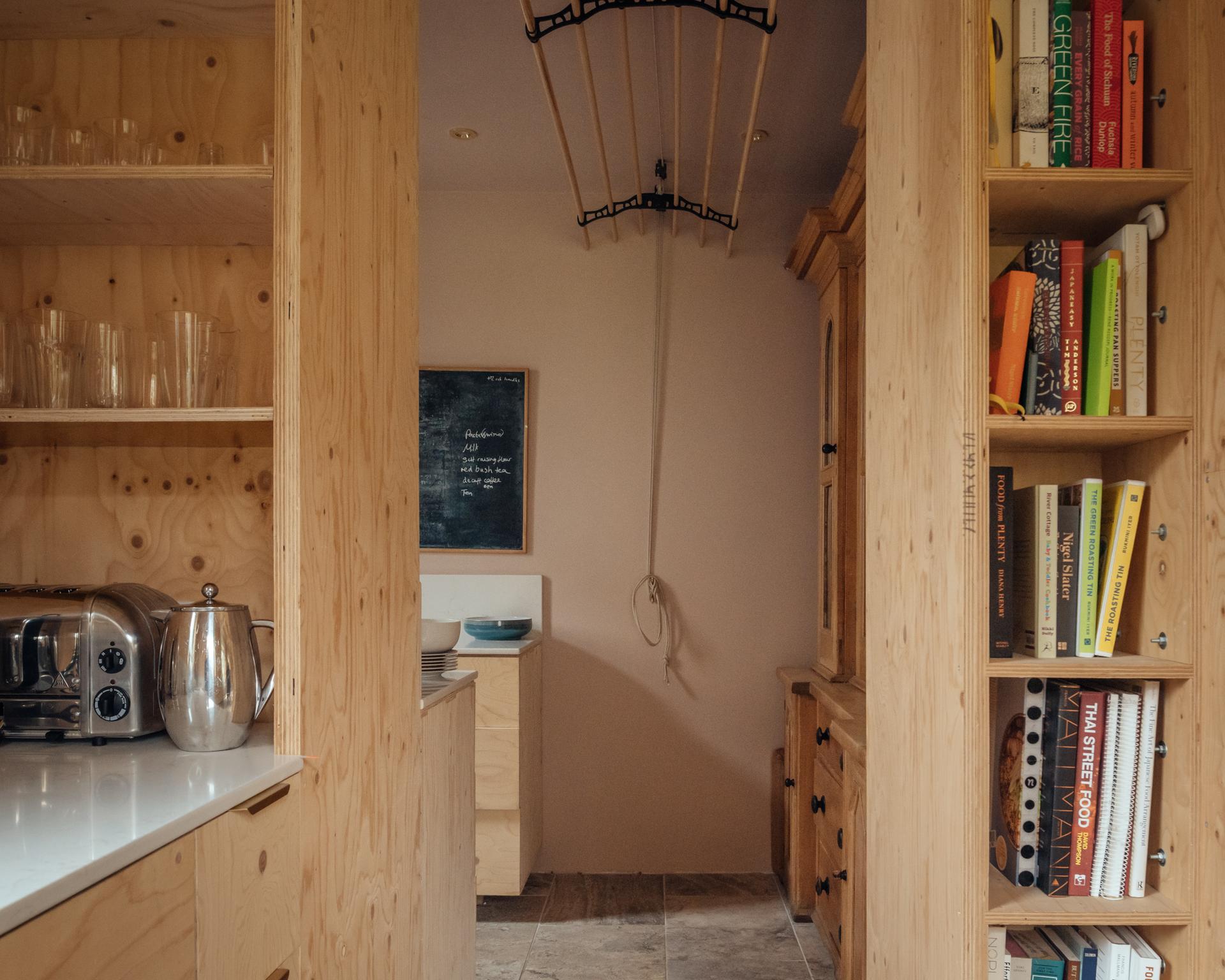 A kitchen featuring timber plywood cabinetry, a toaster and kettle on the left, a built-in bookcase on the right, and a pulley-operated drying rack hanging from the ceiling.