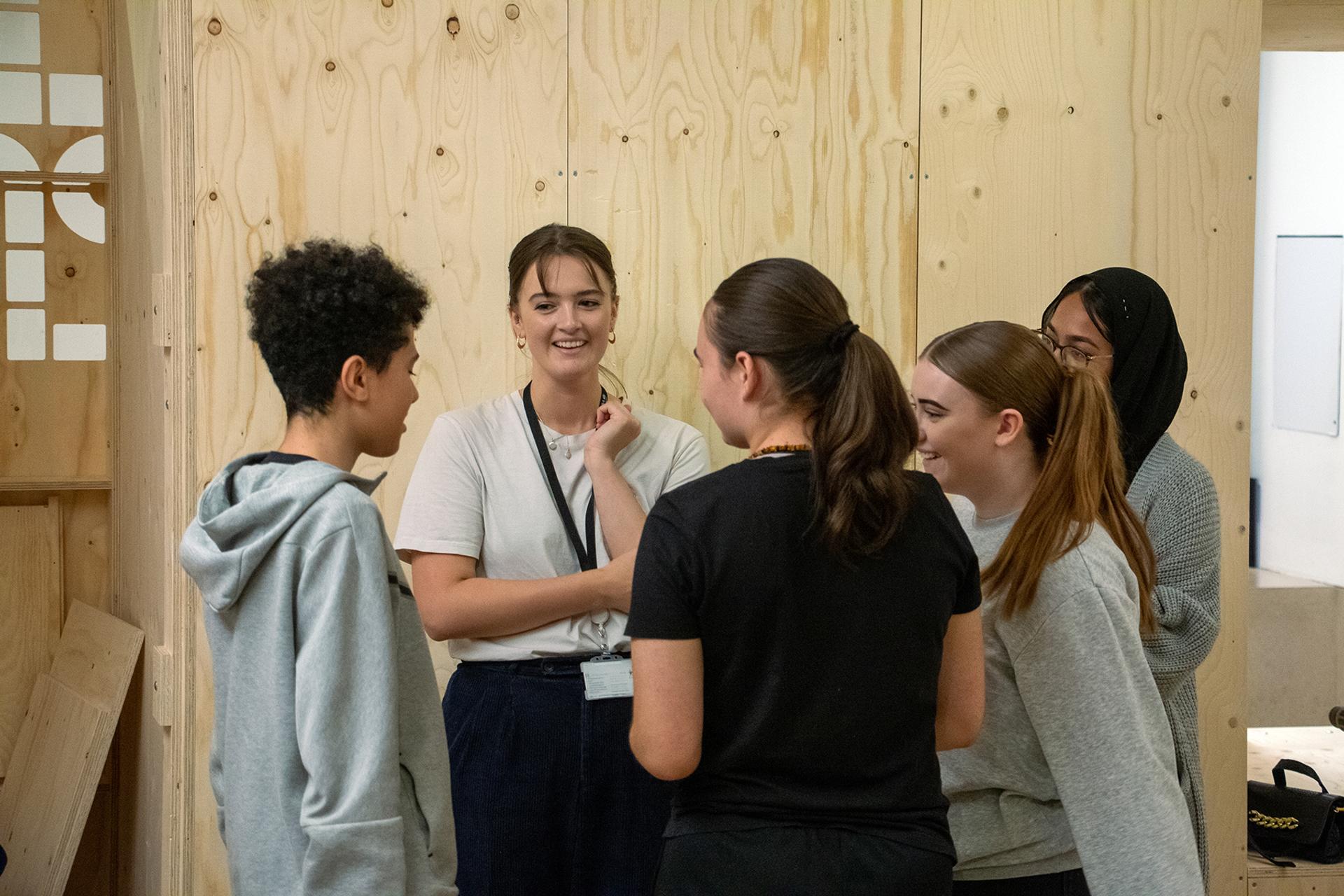 Five young people stand in a circle talking and laughing in a room with birch plywood walls. One person in a white shirt appears to be leading the conversation, whilst others listen and smile.