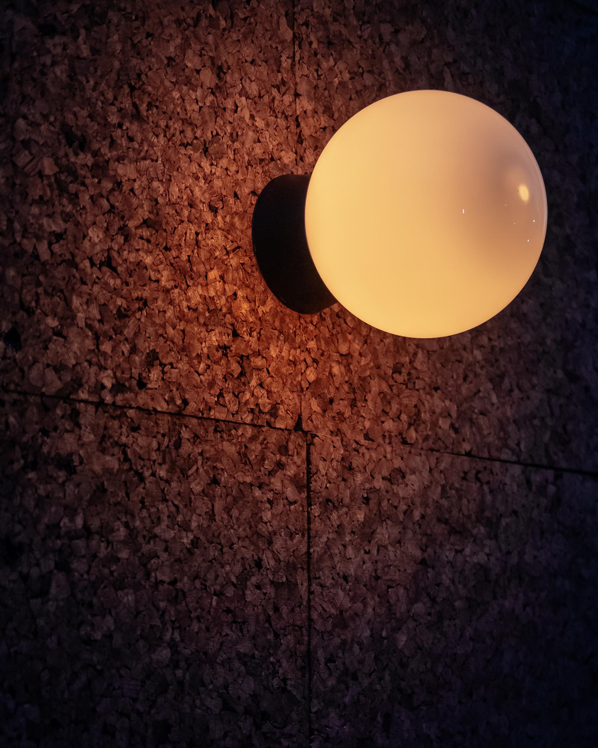 A round, white wall lamp glows warmly against a textured, speckled dark wall, its soft light evoking the natural elegance of birch and plywood often seen in timber construction.