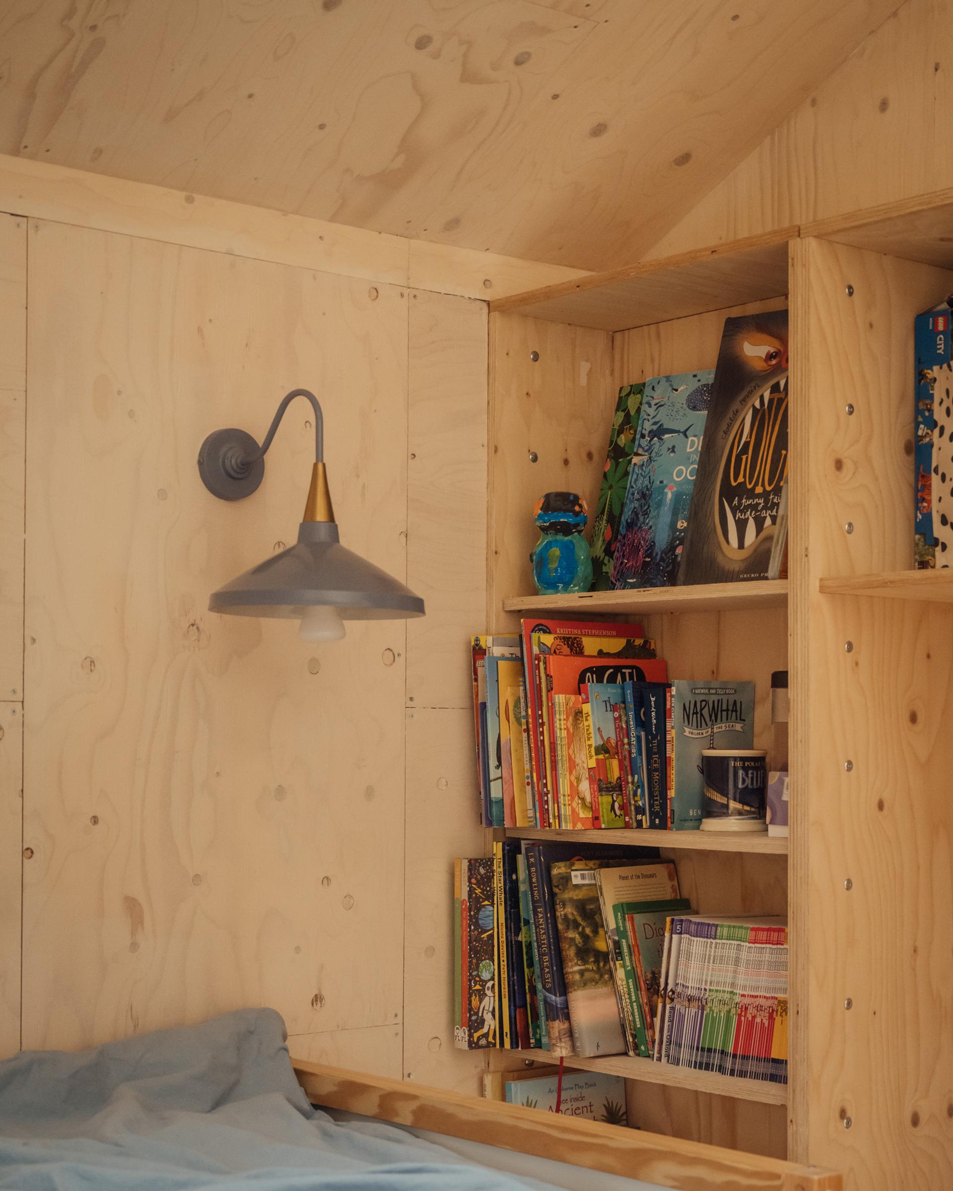 Plywood shelving unit from the U-Build system filled with children’s books, positioned next to a wall-mounted blue lamp above a bed.