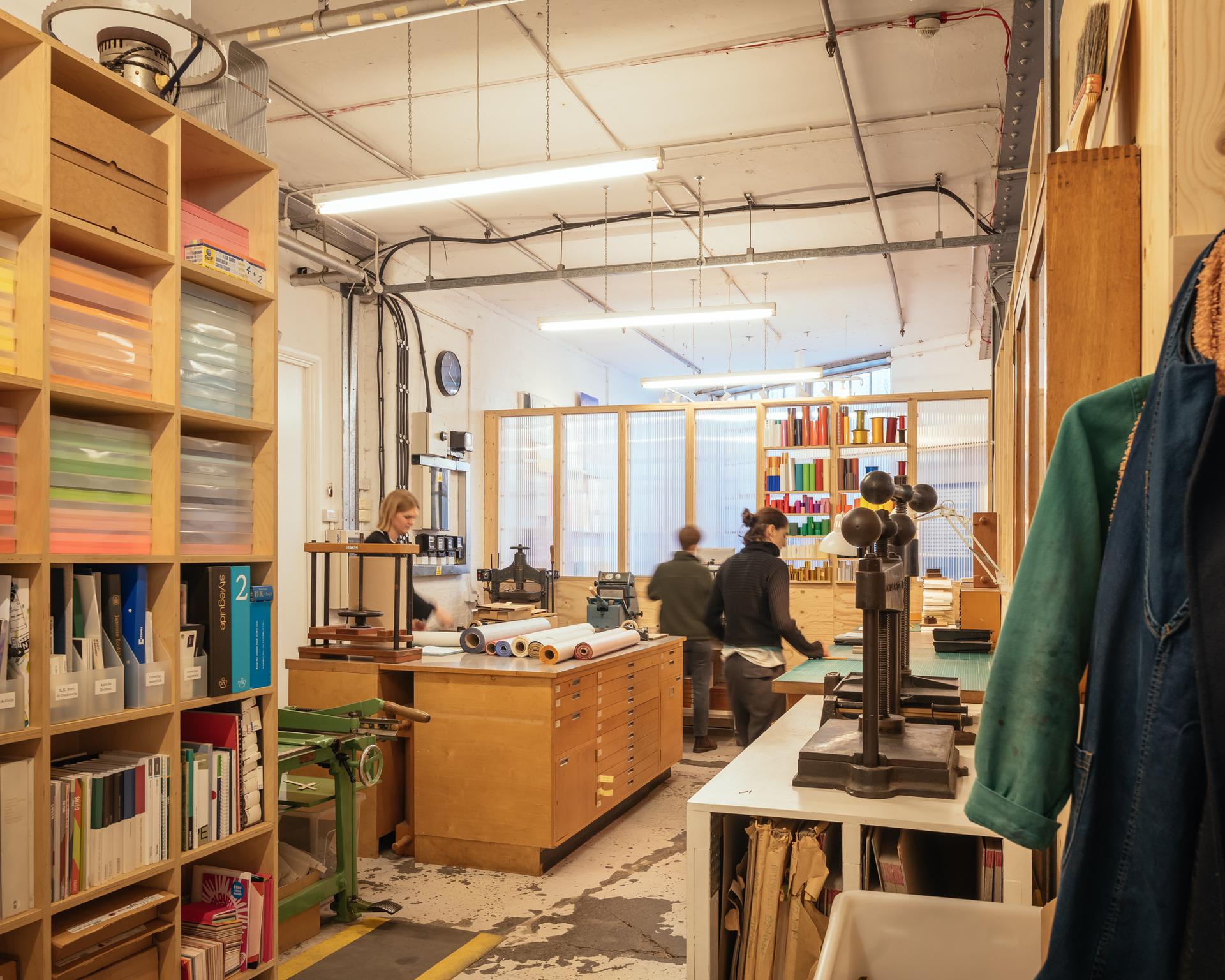 A bright, organised workshop with timber shelves holding papers and books, plywood worktables with tools, and three people working. Rolled papers and colourful materials are visible throughout this sustainable creative space.