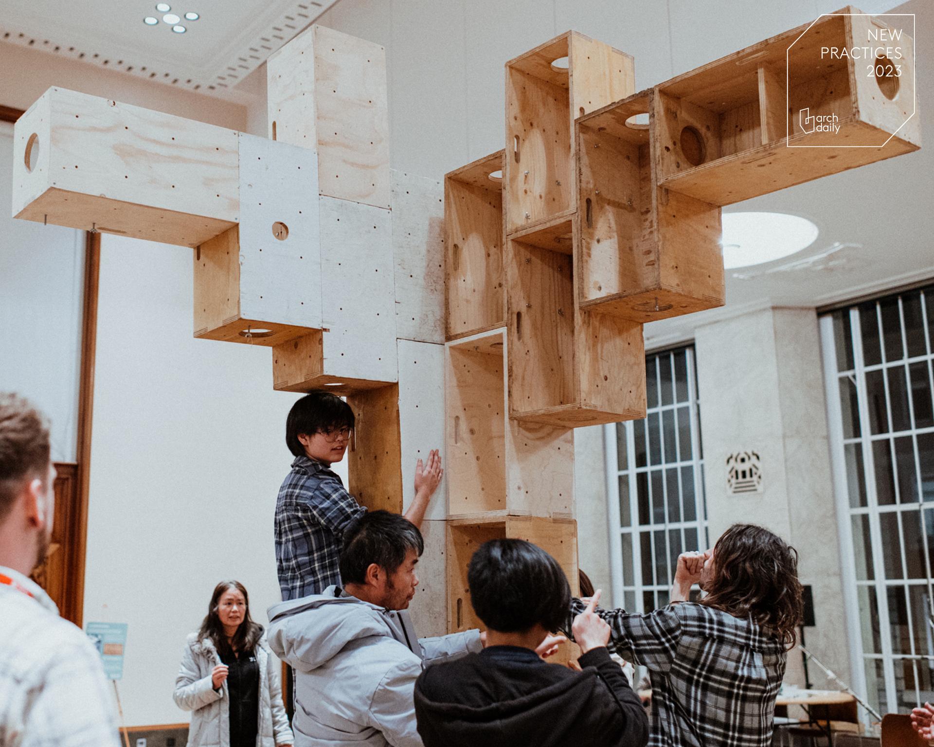 Several people assemble or admire a large birch timber construction indoors, with one person making adjustments whilst others take photos. The atmosphere appears collaborative and creative.