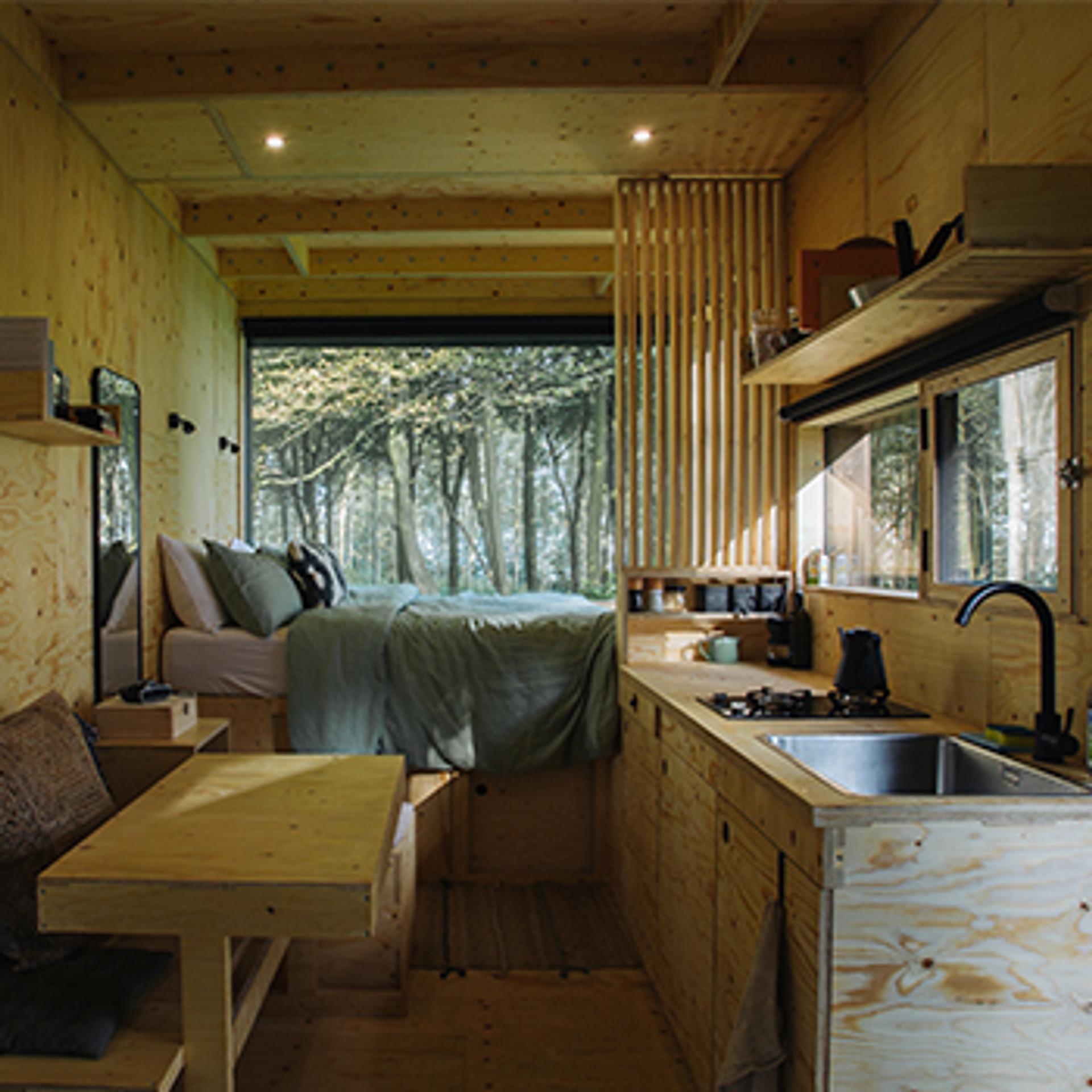 A cosy, modern tiny house interior with wood panelling made from sustainably sourced materials, a compact kitchen, a small dining table, and a bed by a large window overlooking the forest. Natural light fills the space, enhancing its warm and inviting atmosphere.