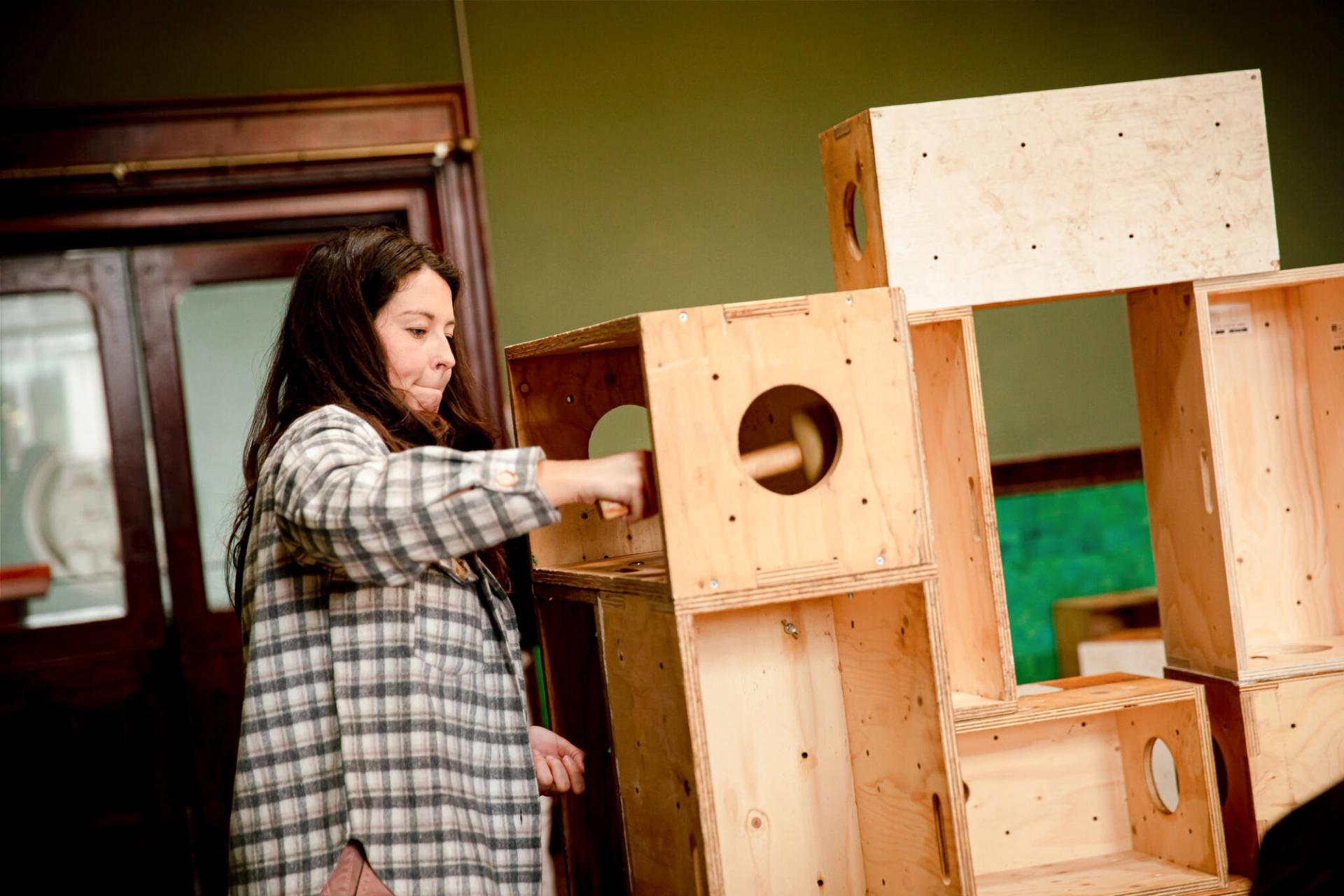 A person in a checked shirt interacts with a stack of plywood boxes with circular holes, possibly building or organising them in an indoor timber construction setting.