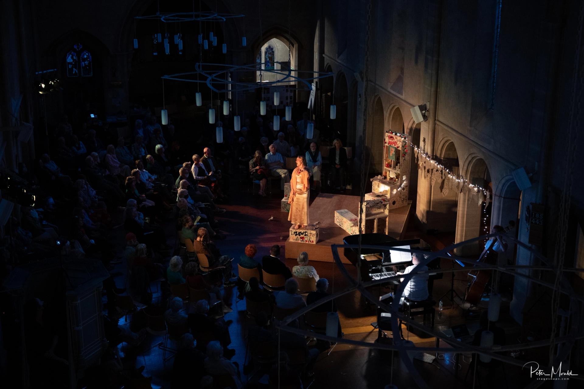 A performer stands under a spotlight on a church stage with timber construction, surrounded by an audience in a semicircle. Musicians play nearby as hanging lights softly illuminate stained glass windows in the spruce-framed background.