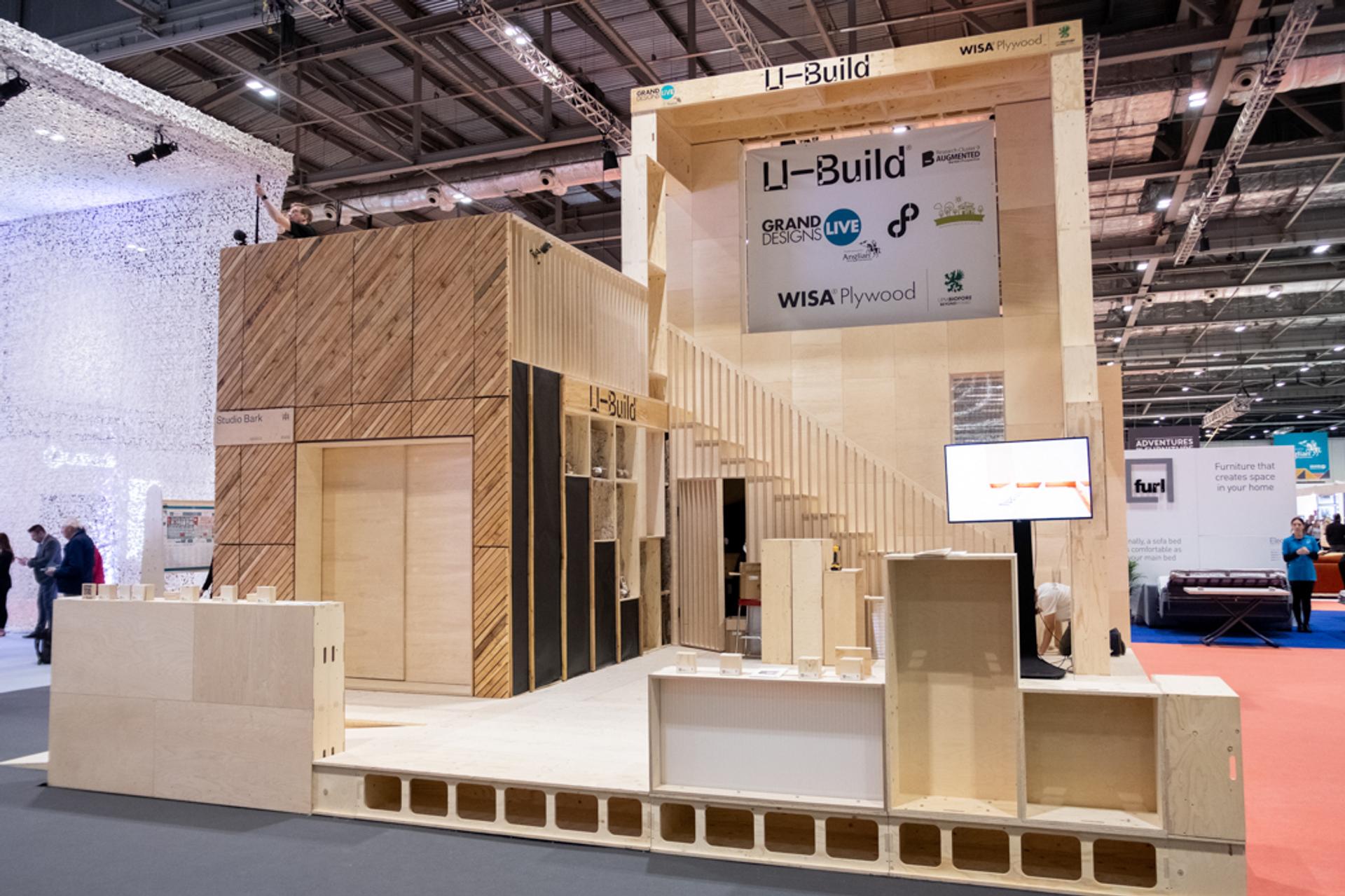 A modern, large wooden exhibition stand crafted from spruce with geometric patterns, signage reading U-Build, and display screens, set up in a spacious, well-lit event hall with other stands visible in the background.