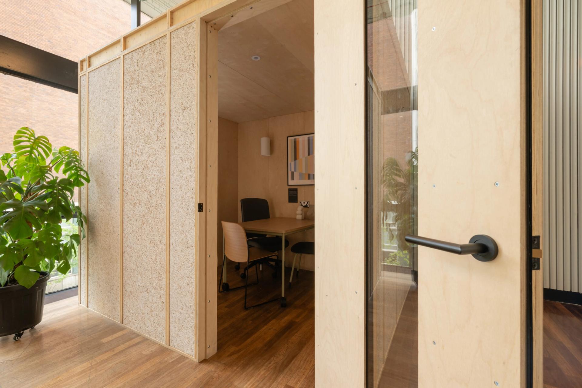 Small modern meeting room with wooden walls, a glass door, two chairs at a table, a plant outside, and a painting on the wall—all flooded with natural light—resembling the stylish Meeting Pods found in the Renold Building.