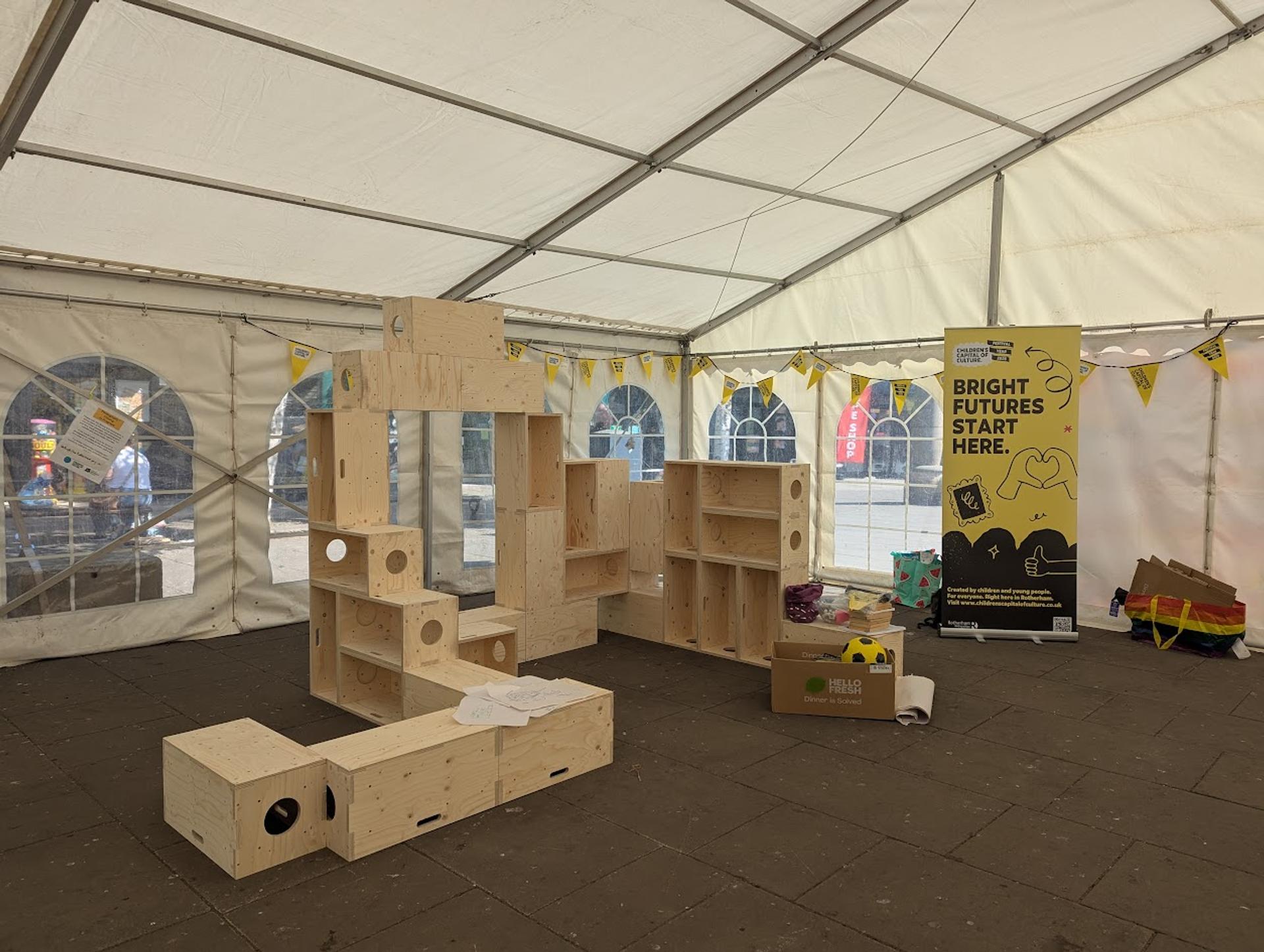 Wooden birch modular play structures are assembled inside a white marquee. A colourful display and a large sign reading Bright Futures Start Here stand out, with bunting flags hanging along the marquee walls.