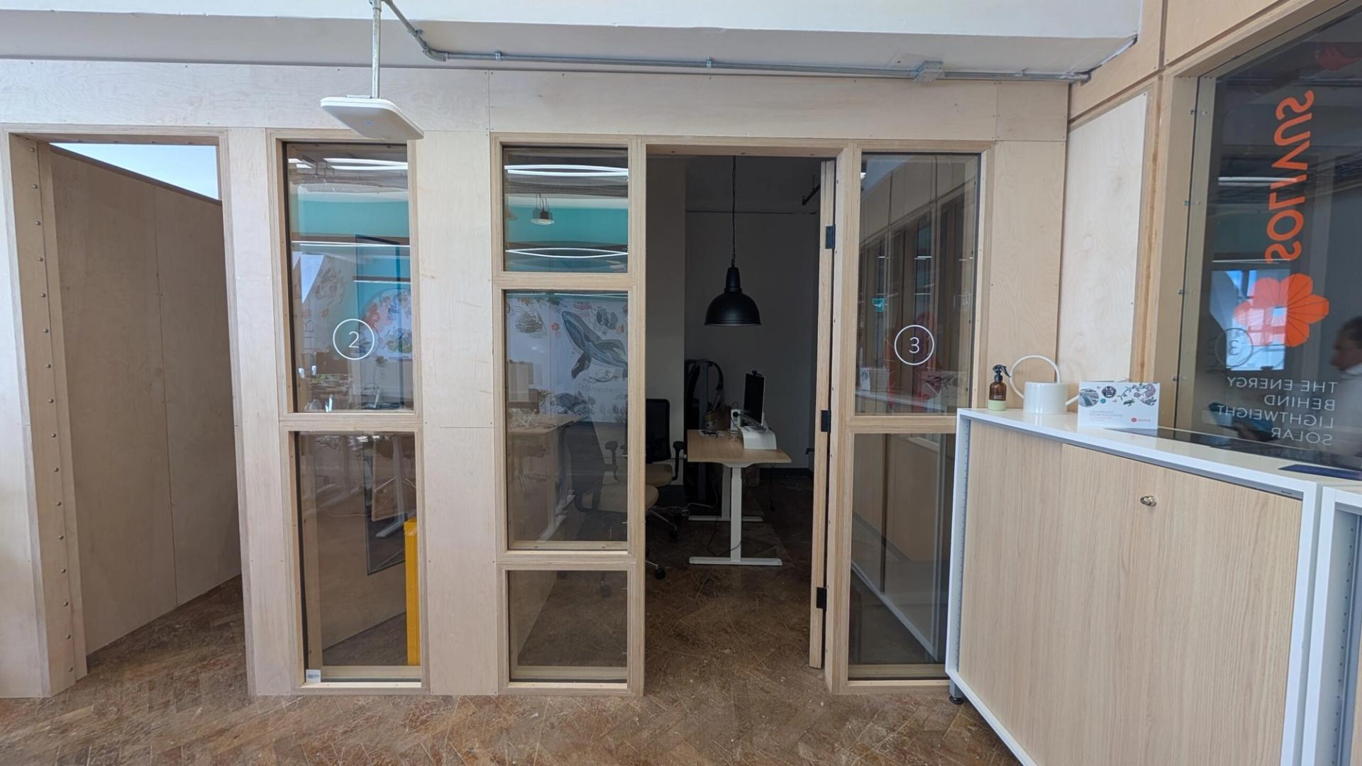 Small modern office space with plywood walls and glass doors labelled “2” and “3.” Inside, a desk, computers, chair, and black lamp are visible. A white cupboard and potted plant sit outside office number 3.