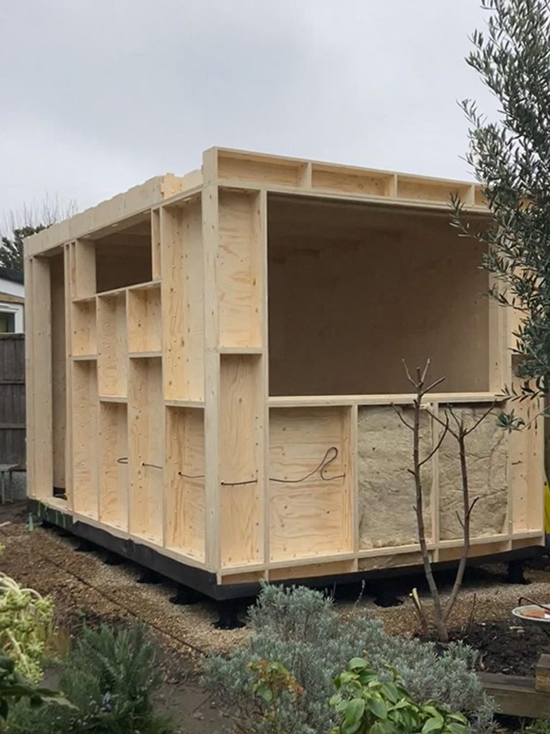 A partially constructed modular wooden shed stands in a garden, its spruce framing and unfinished open wall exposed among various plants on a cloudy day.