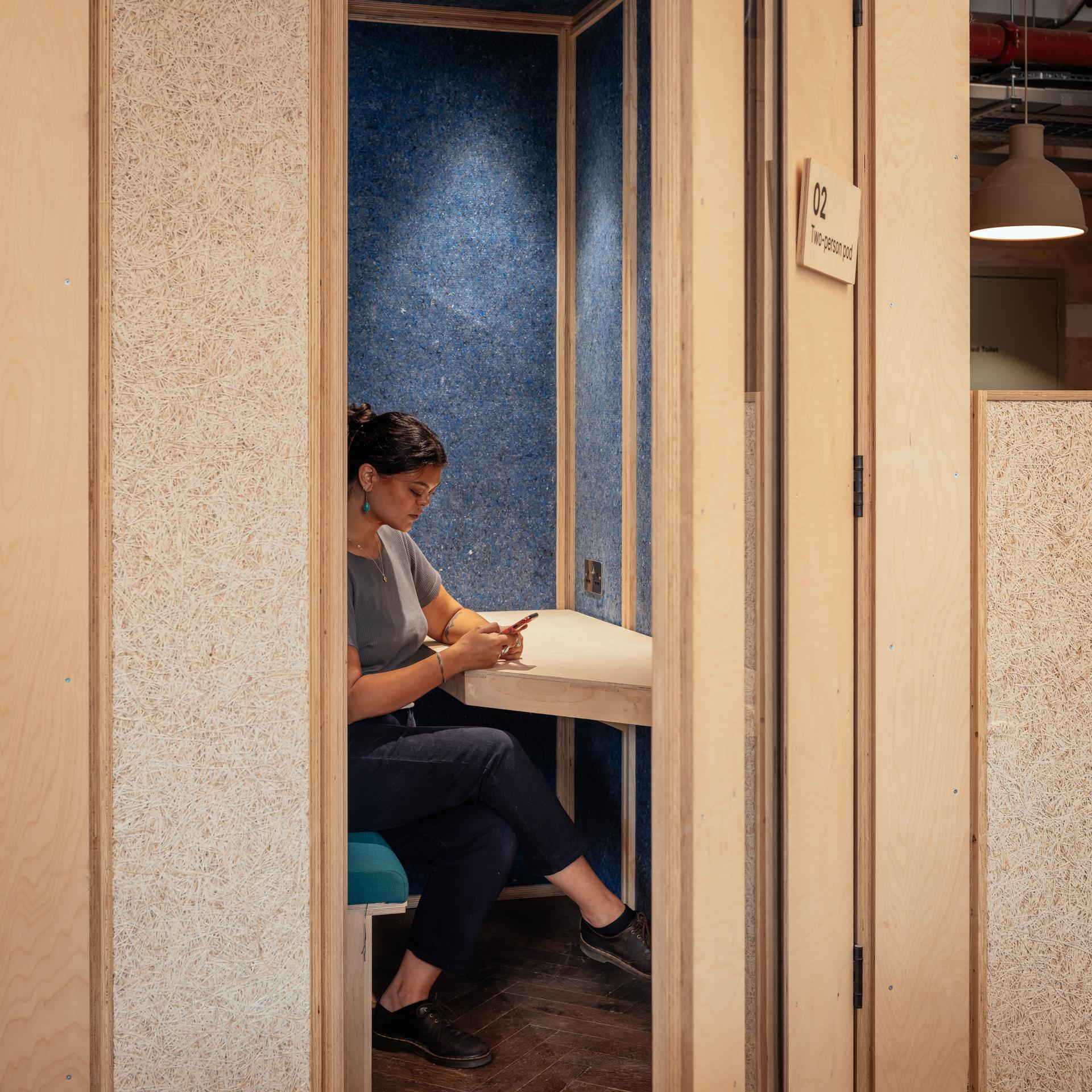 A woman sits alone in a small, private cubicle with a blue interior and spruce timber construction. She looks at her mobile at a small desk, surrounded by light wood panels. A sign outside the cubicle reads A02.