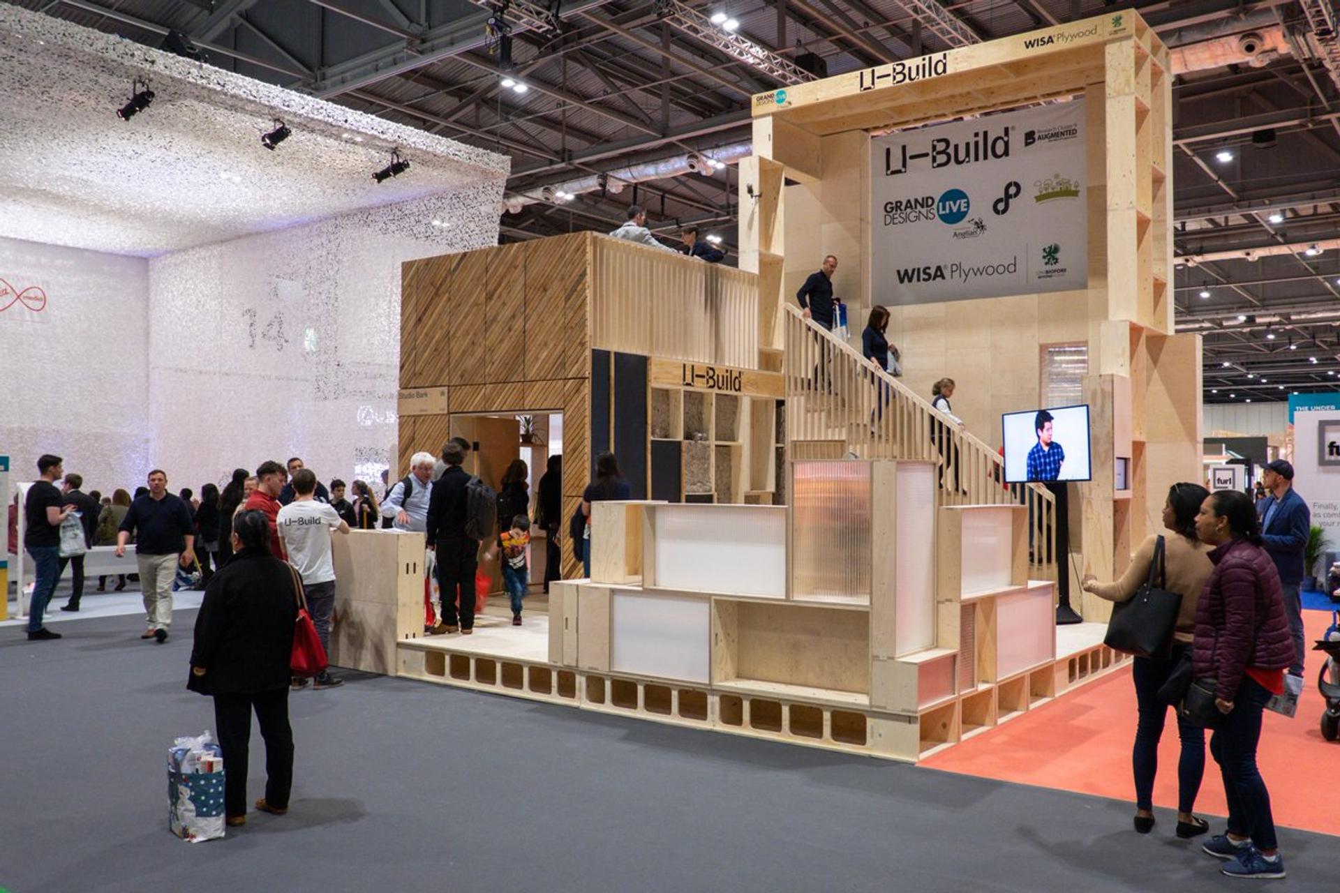 A busy exhibition hall with people gathering around a large modular timber construction trade show stand for Li-Build, featuring birch stairs, signage, and displays under bright overhead lighting.