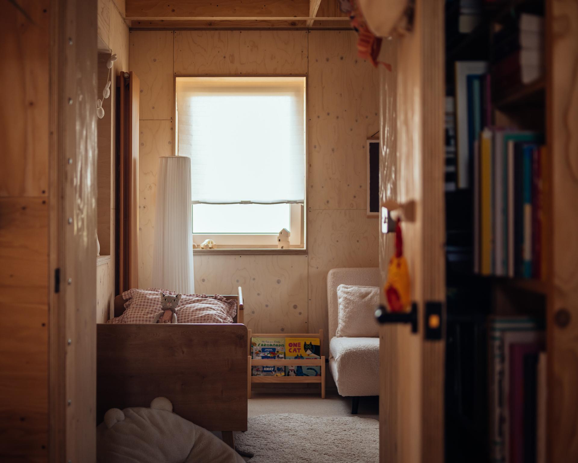 A cosy child’s room with plywood walls, a small wooden bed, a low bookcase, and a textured armchair, viewed through the doorway of a home extension.