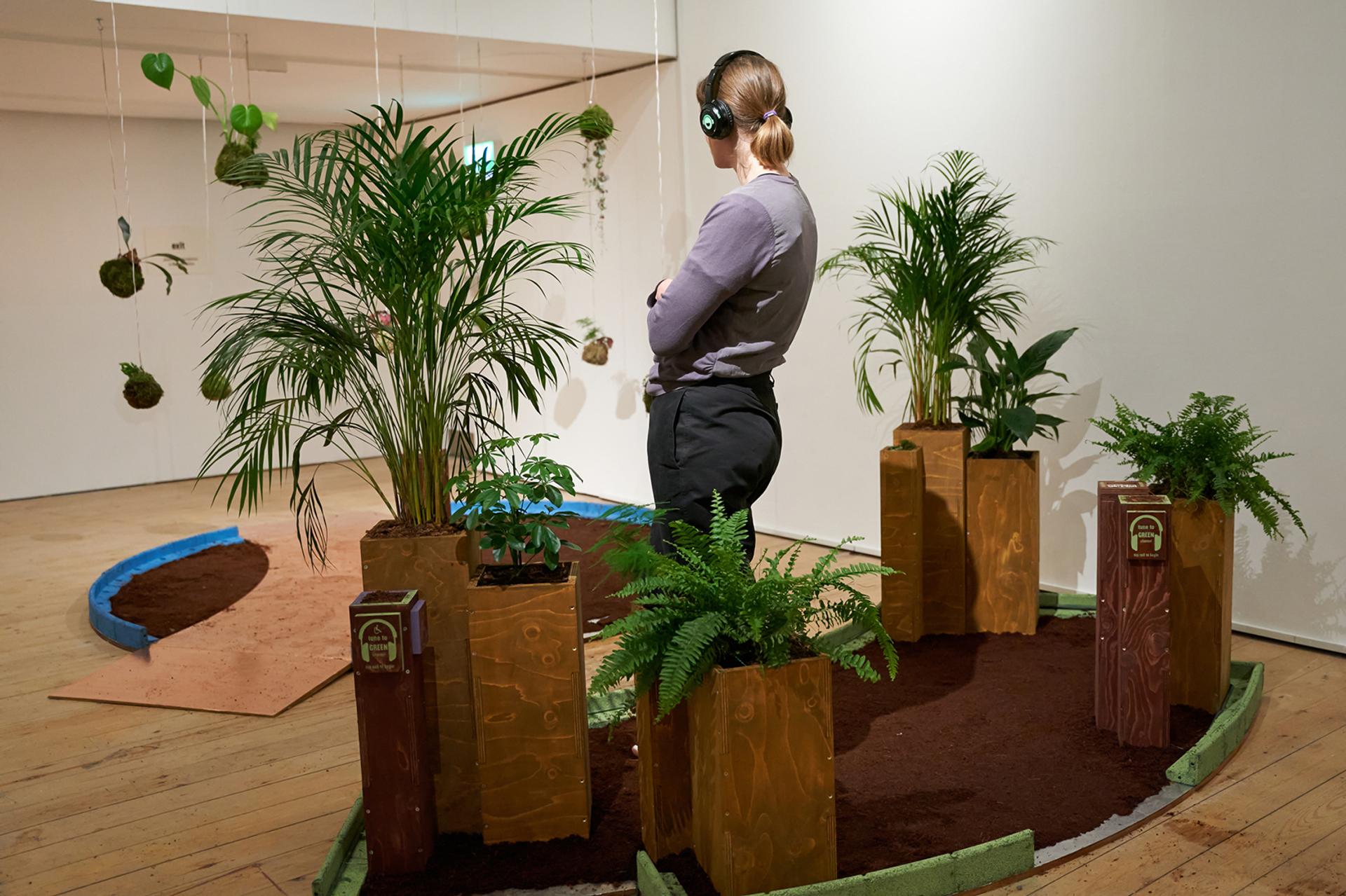 A person wearing headphones stands among potted plants on a soil-covered floor in an indoor exhibition space, surrounded by decorative plywood boxes and some hanging plants—a nod to sustainable design.