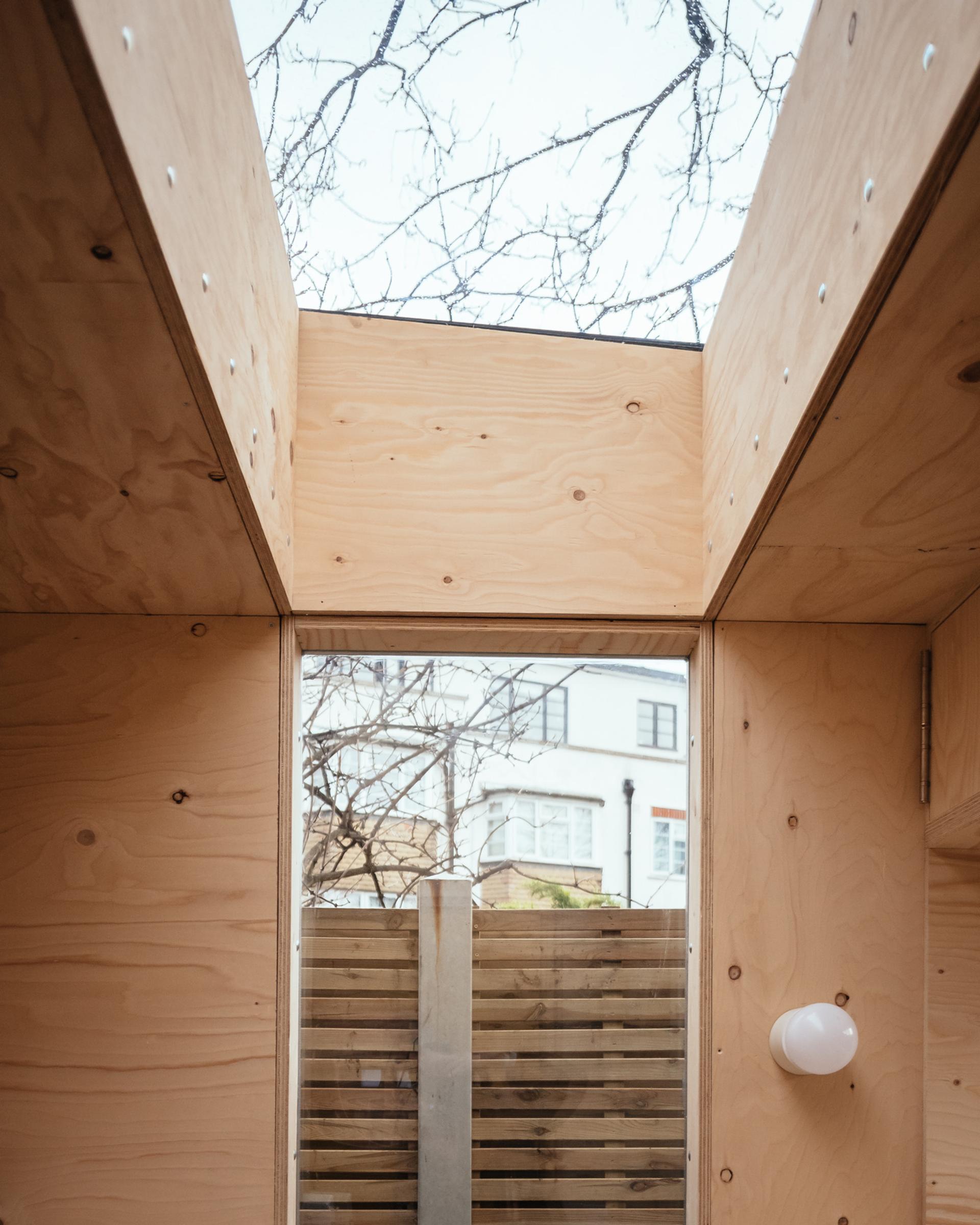A wooden interior with light-coloured, sustainably sourced plywood walls features a skylight above and a glass door leading to a fenced outdoor area; tree branches and a white building are visible outside.