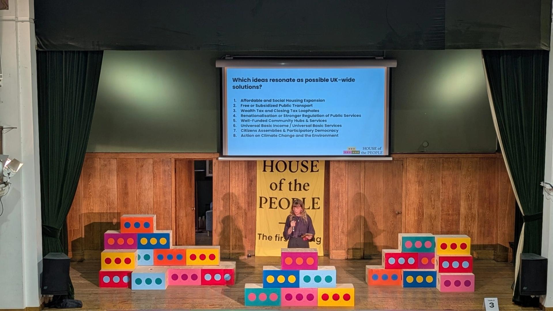 A speaker stands on stage in front of colourful, upcycled building blocks and a sign reading HOUSE OF THE PEOPLE, presenting a slide about possible UK-wide solutions to an audience in a wood-panelled hall.