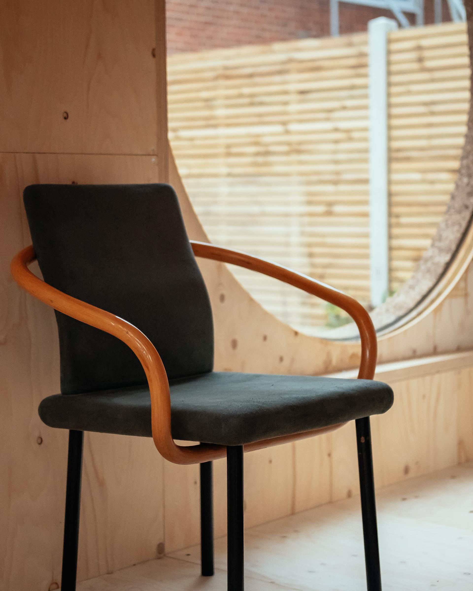 A modern chair with a dark cushion and curved wooden armrests—crafted with sustainability in mind—sits in front of a large circular window, with a wooden fence and brick wall visible outside.