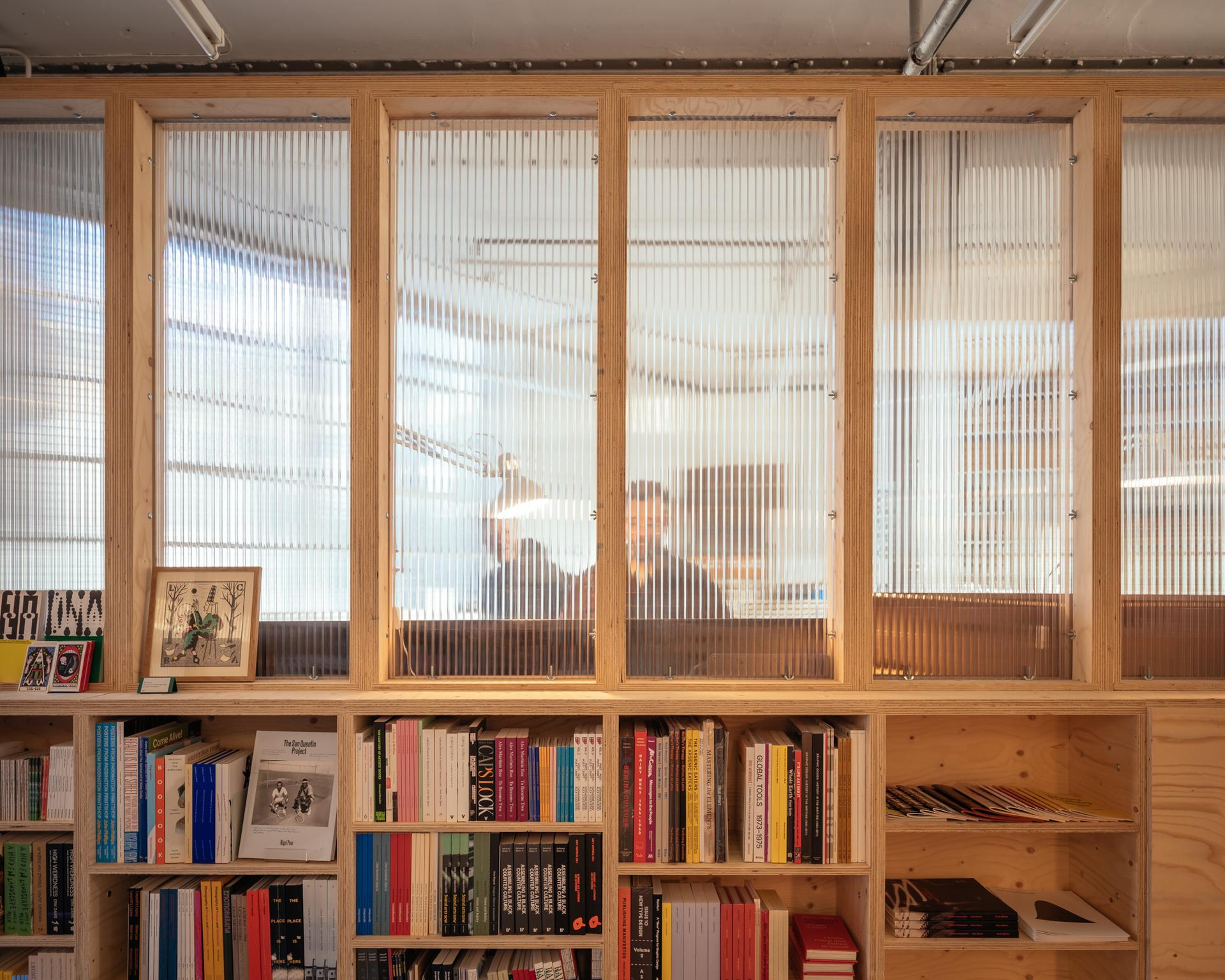 Bookshelves filled with colourful books sit below tall, translucent birch panels that partially obscure two people working at desks, with soft, natural light filtering into the room.