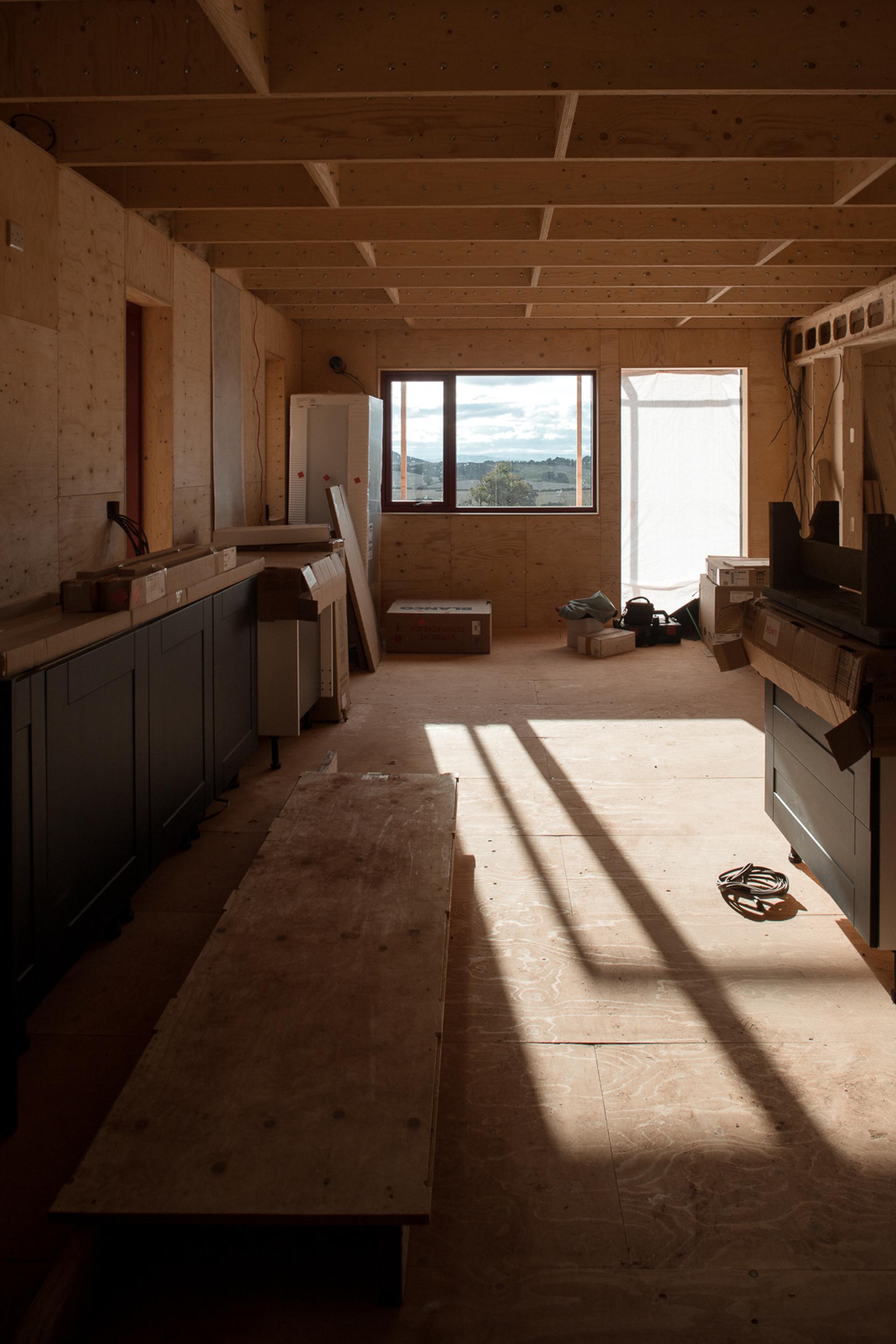 A partially constructed room with unfinished wooden floors and walls made from sustainably sourced timber, scattered tools and materials, and sunlight streaming through a window, casting long shadows across the floor.
