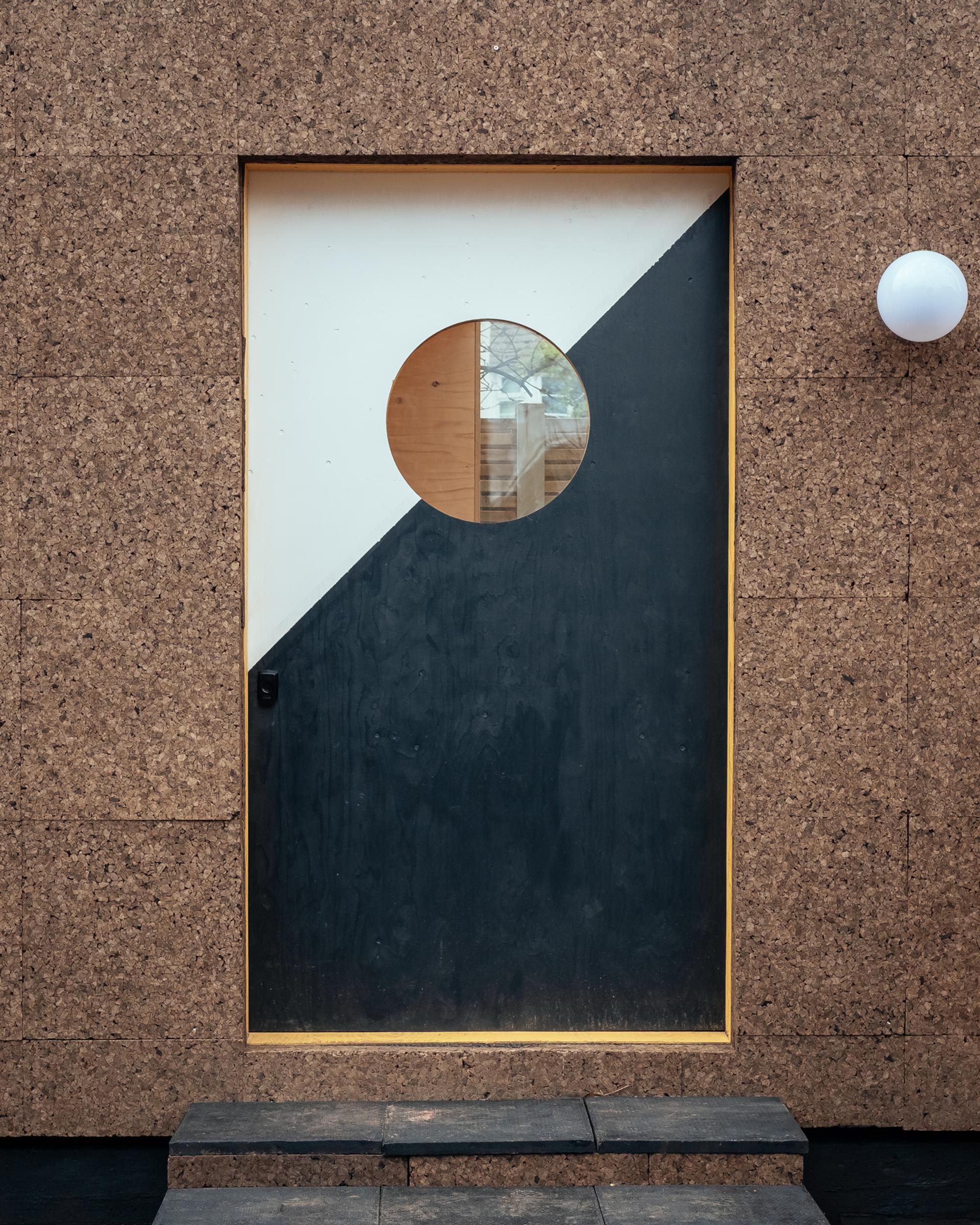 A modern door with a diagonal black and white design and a round window, set in a birch plywood-textured wall. A white spherical light fitting is mounted on the modular wall to the right of the door.