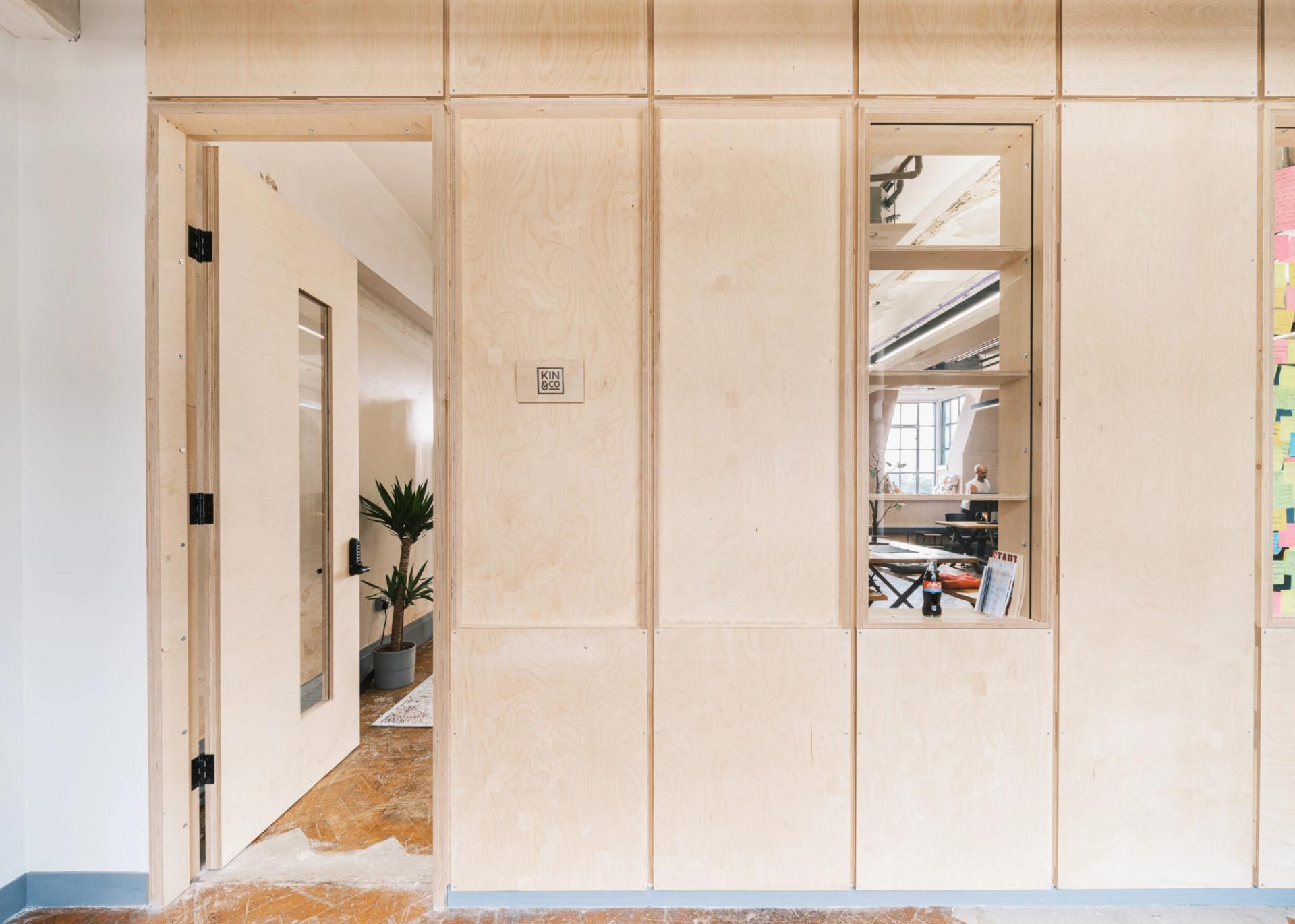 A modern office with light spruce wood panelling, an open door, an indoor plant, and windows revealing a bright workspace with people and desks inside.