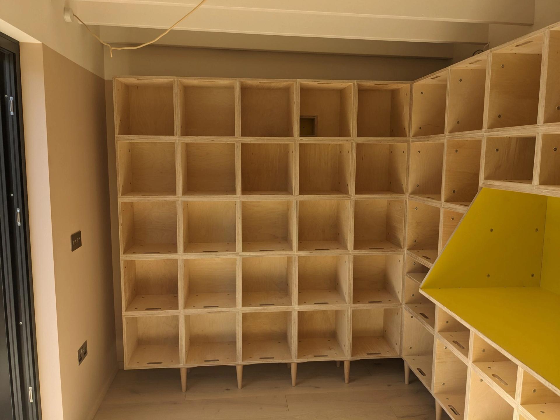 Wooden shelving unit with multiple square cubbyholes fills two walls of this East London Record Room. One section includes a shelf with a bright yellow interior. The room has light walls, a light wooden floor, and exposed ceiling beams.