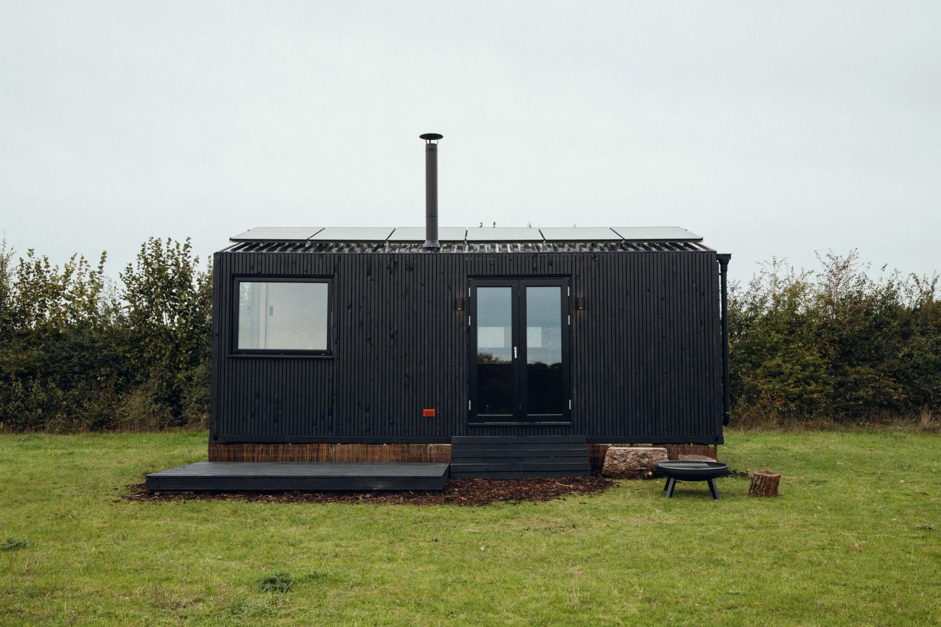 A small black modular tiny house with solar panels on the roof, a central door, and a large window sits on a grassy field with bushes in the background. A fire pit, chair, and logs enhance this sustainable retreat.