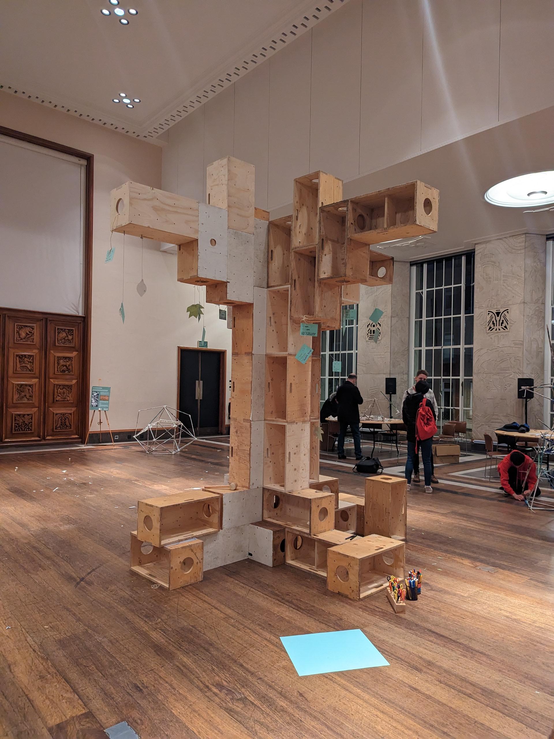 A large modular, spruce tree-like structure made of stacked boxes stands in a spacious indoor room with high ceilings and wooden floors. People are nearby, and various papers and objects are scattered on the floor.