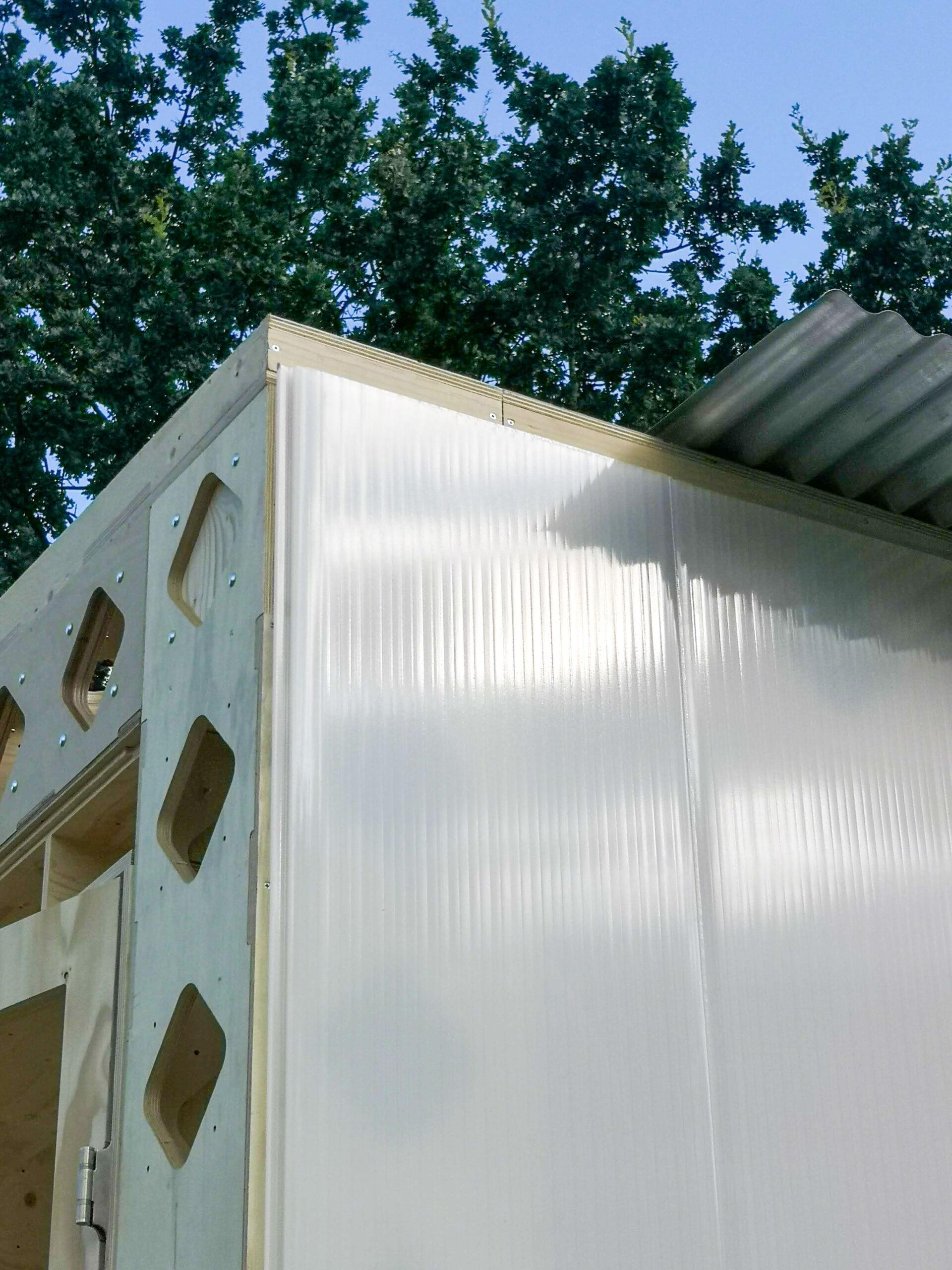 A close-up of a modern building corner featuring translucent corrugated panels and light-coloured spruce plywood, set against a backdrop of green leafy trees and blue sky.