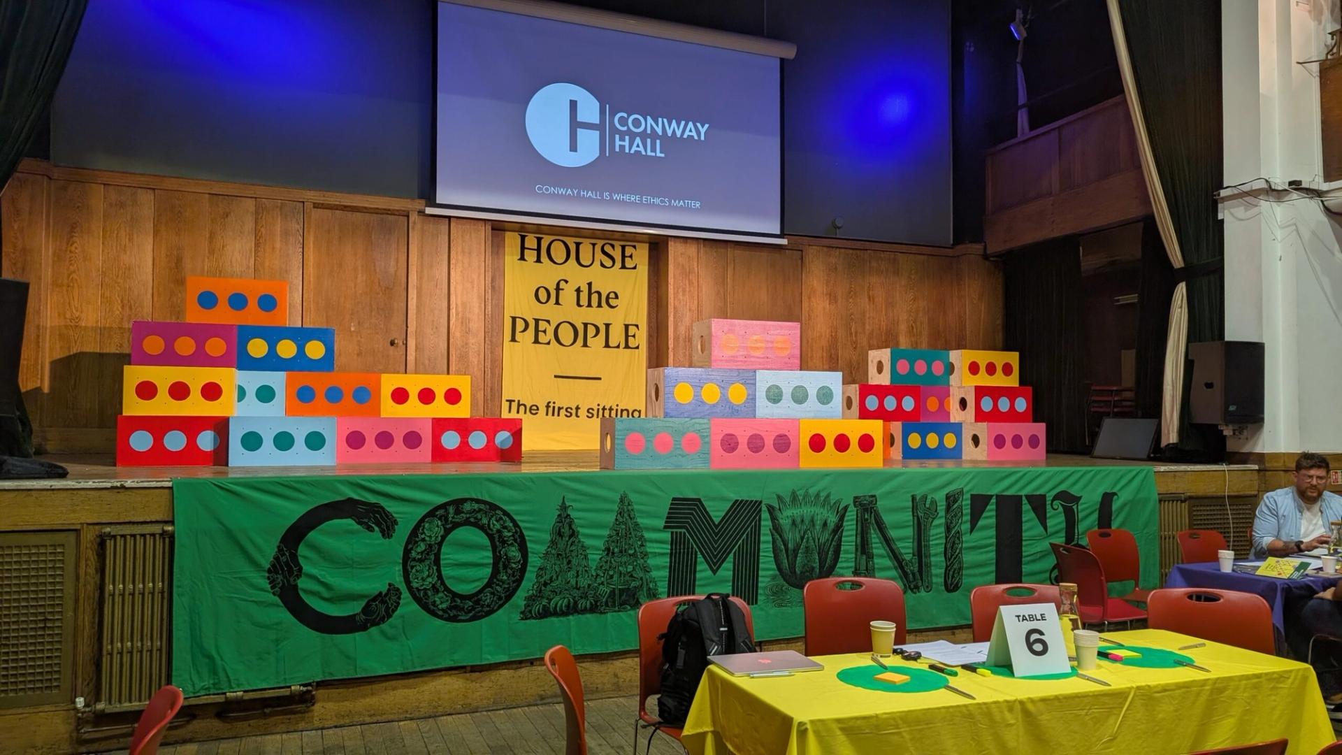 A decorated hall with colourful brick props, a green COMMUNITY banner, and a House of the People sign. The screen displays Conway Hall. Red chairs and tables, made from sustainably sourced materials, are set up; one table is labelled Table 6.