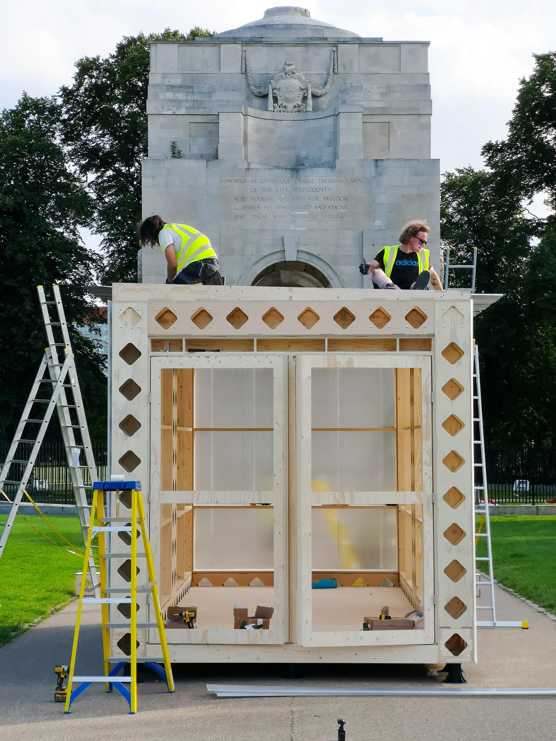 Two people in yellow safety vests work on the roof of a sustainable wooden structure under construction, built with birch plywood, in front of a large stone monument. Ladders and tools surround the site.