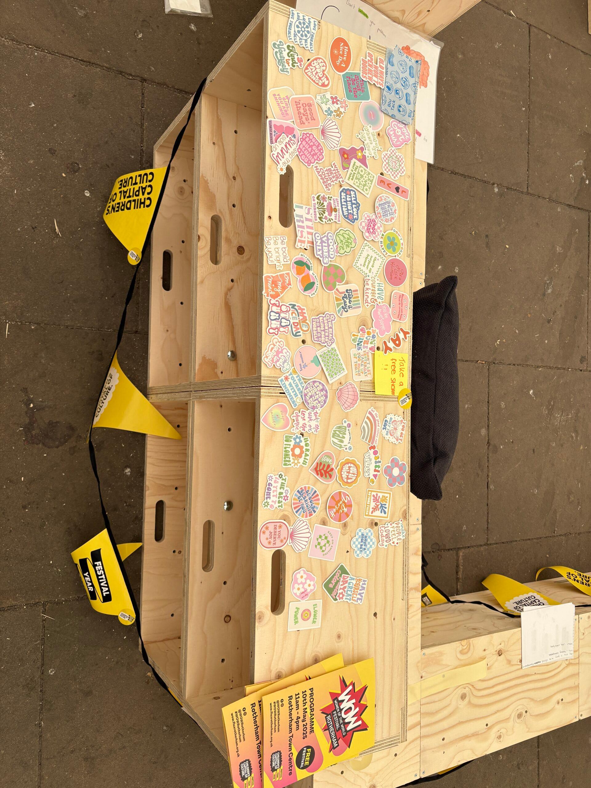 A birch plywood bench displays colourful, hand-drawn stickers and decorations. Yellow signs nearby read Children's Capital of Culture. The scene appears to be part of a creative, sustainable event or workshop.