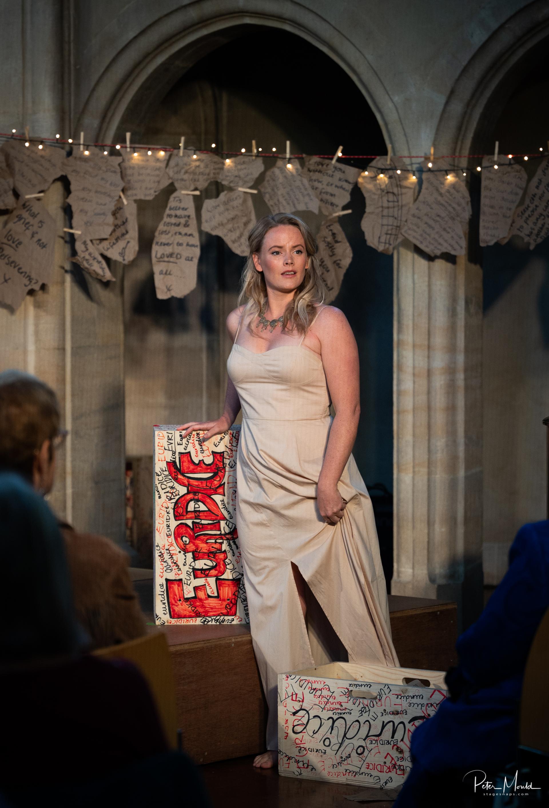 A barefoot woman in a beige dress stands on a stage, holding a painted sign. Audience members watch her. Behind her, fairy lights and handwritten notes hang in front of arched stone columns built with sustainable timber construction.