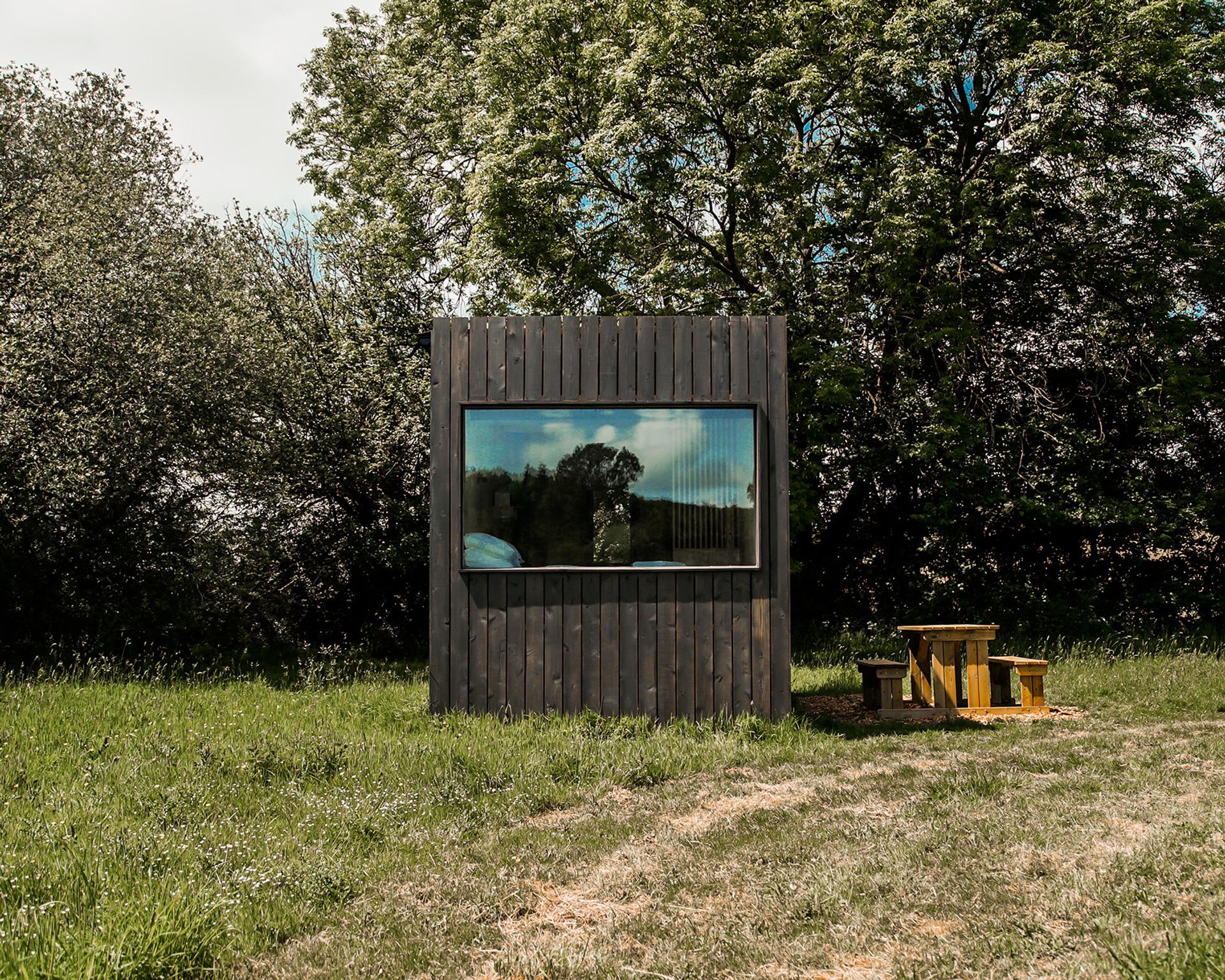 A small, modern wooden cabin built from sustainably sourced materials stands in a grassy field with trees behind. A picnic table with benches sits nearby, and the cabin’s large window reflects the surrounding greenery and sky.