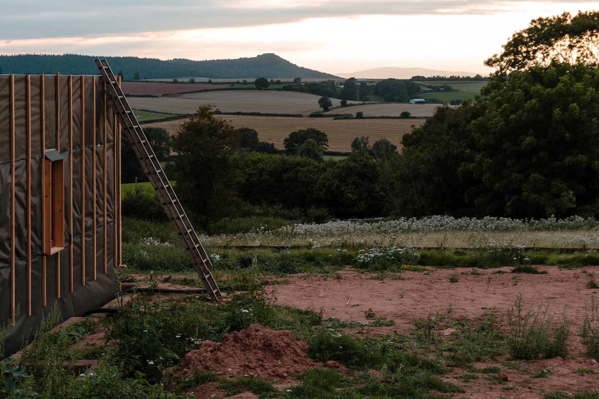 A building site with a ladder leaning against a partially built, upcycled structure, surrounded by earth and wildflowers, overlooks rolling fields and distant hills at sunset.